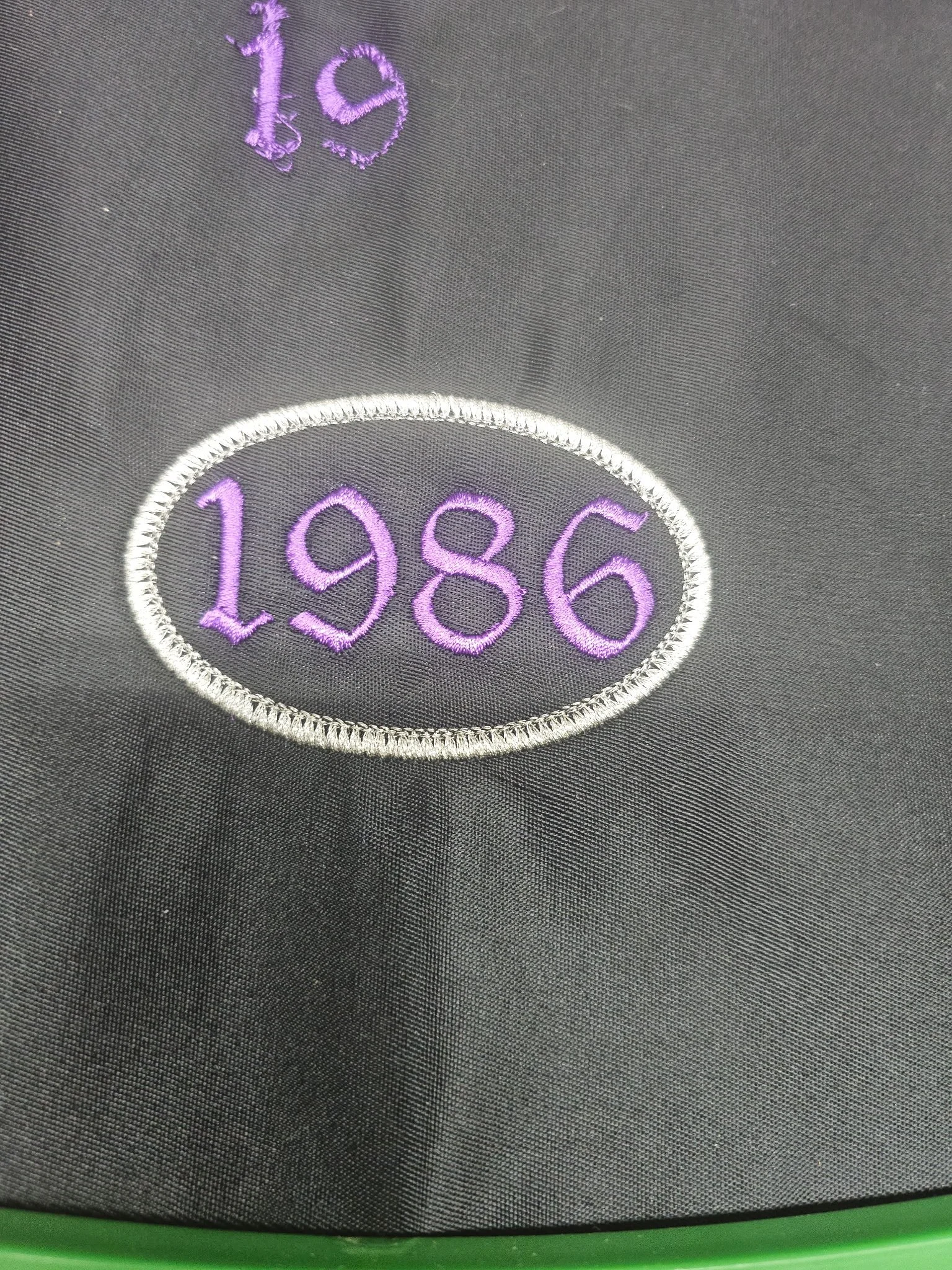 Close-up of black fabric with embroidered purple numbers '1986' inside a white stitched oval, and the number '19' partially visible embroidered in purple above.