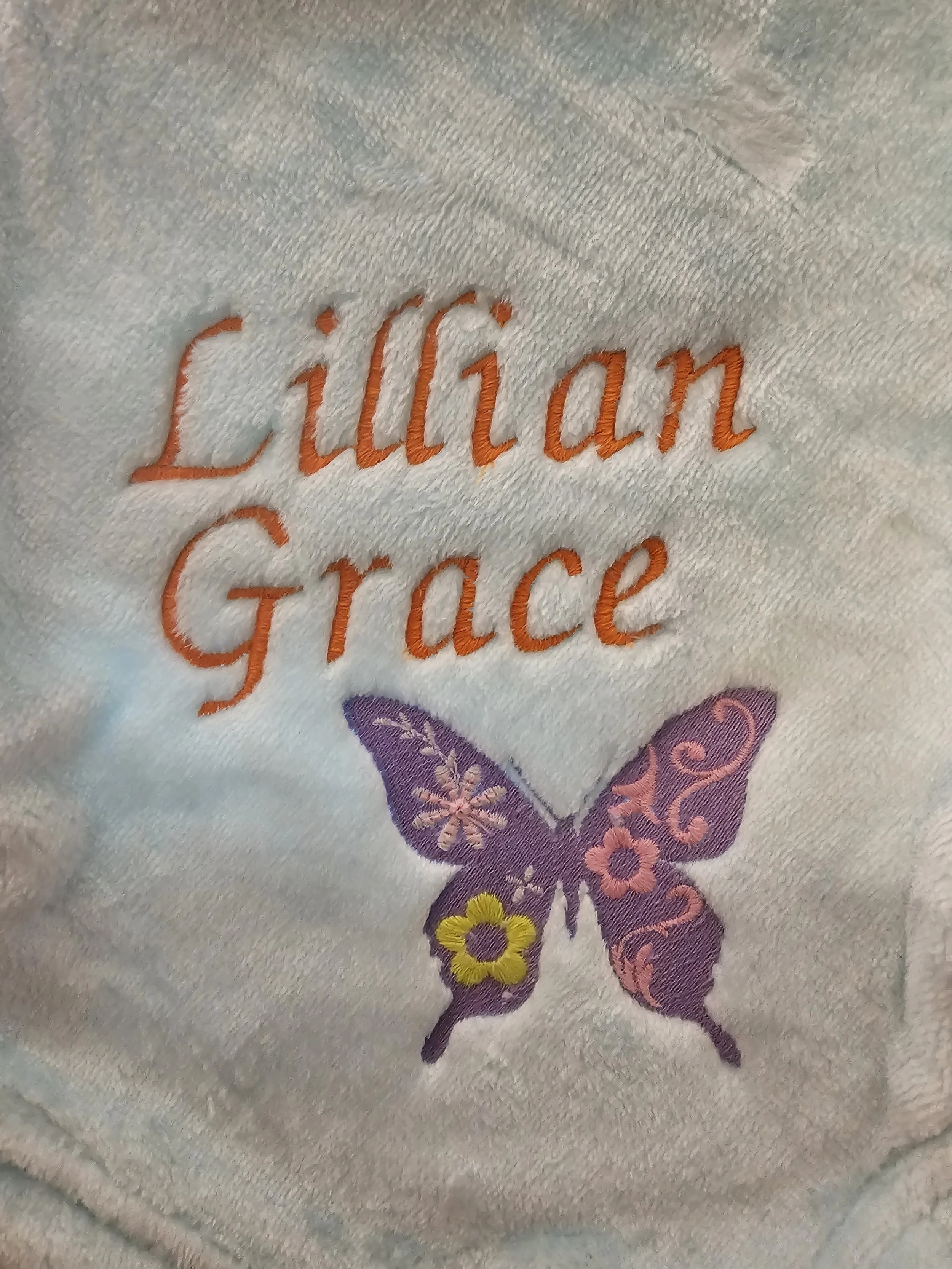 Embroidered label with the words 'Lilies and Grace' in orange thread and a purple butterfly with pink and yellow floral details on embroidered fabric.