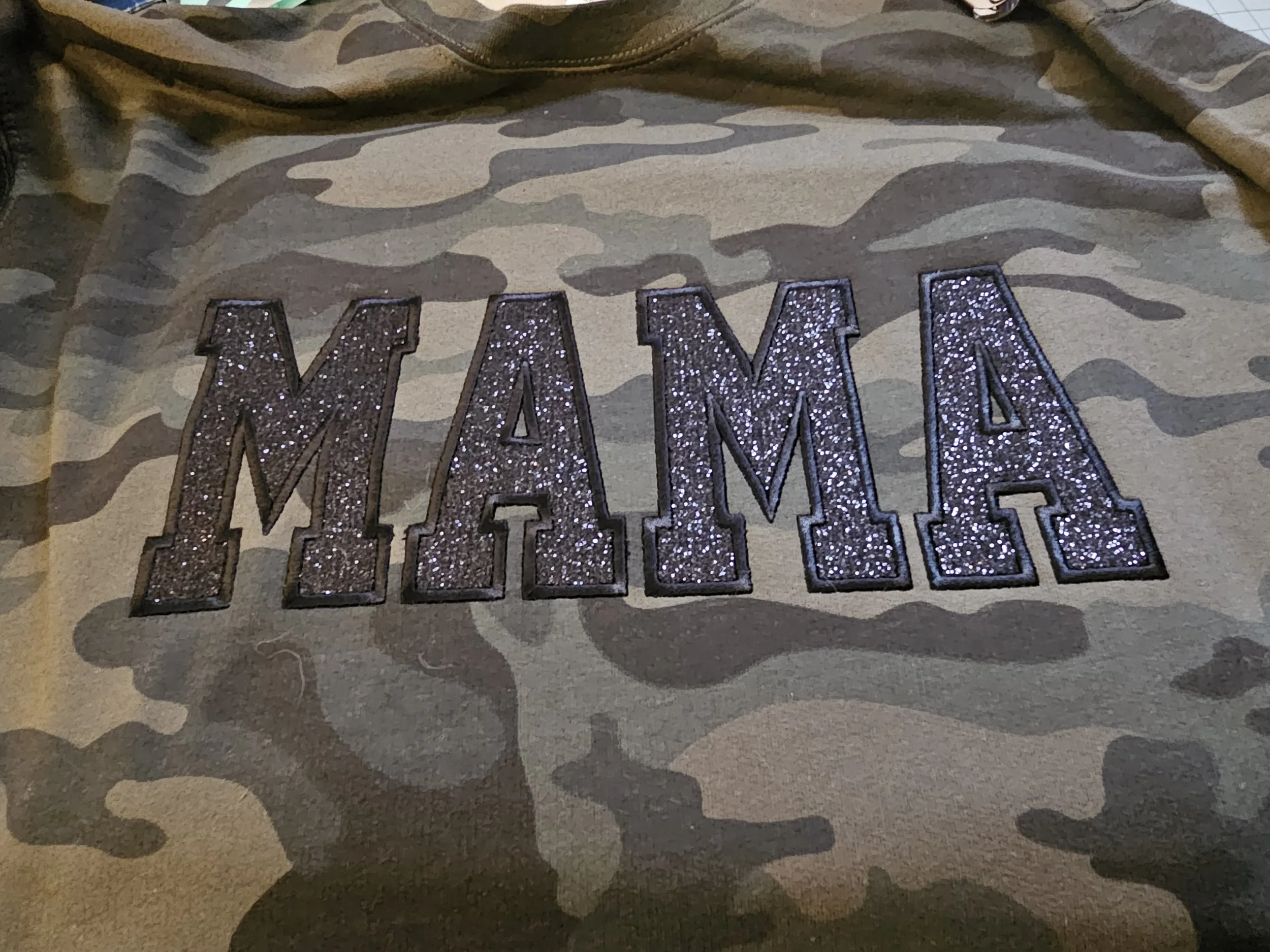 A gray camouflage shirt with the word 'MAMA' in glittery black letters.