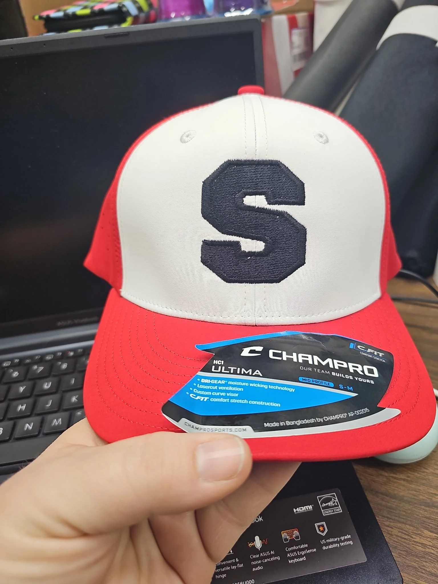 Person holding a red and white trucker cap with a large black 'S' logo on the front, placed on a desk with a computer keyboard and monitor in the background.