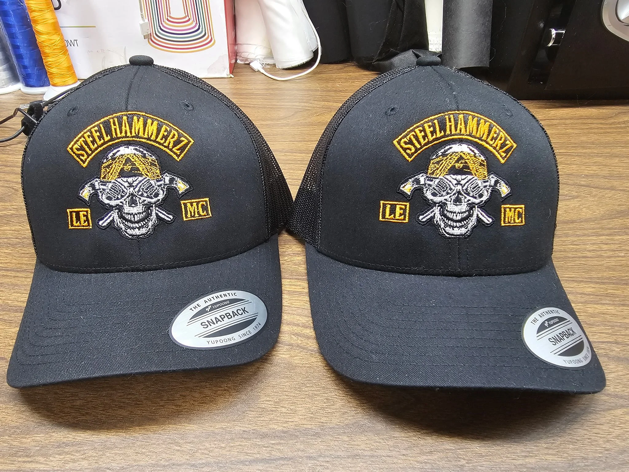 Two black baseball caps with a skull logo, sunglasses, and crossed bones, embroidered with 'Steelhammerz LE MC' on the front, on a wooden surface.