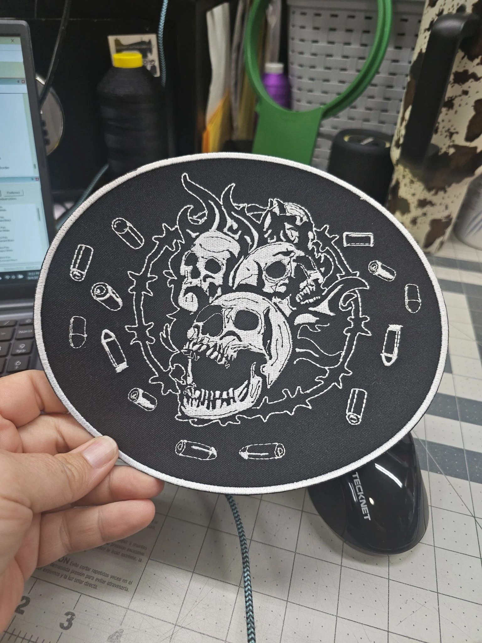 A black patch with white embroidered skulls, flames, and bullets design, held in a person's hand.