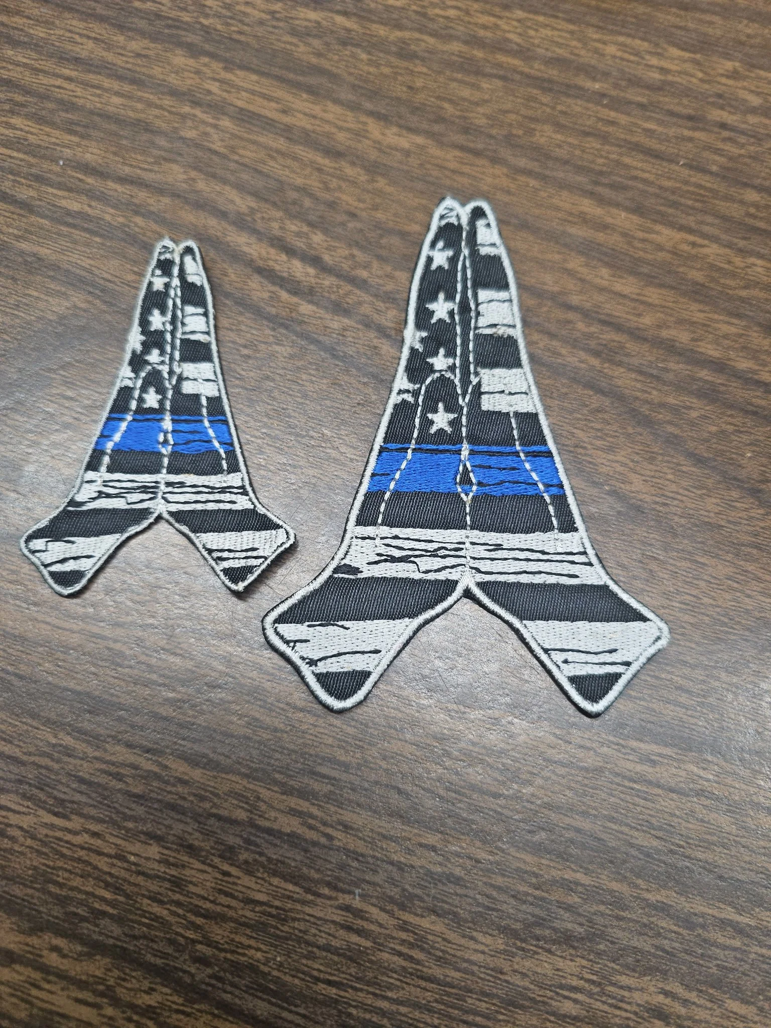 Two embroidered patch patches resembling military or police insignia with a black and white American flag pattern and a single blue stripe, laid on a wooden surface.