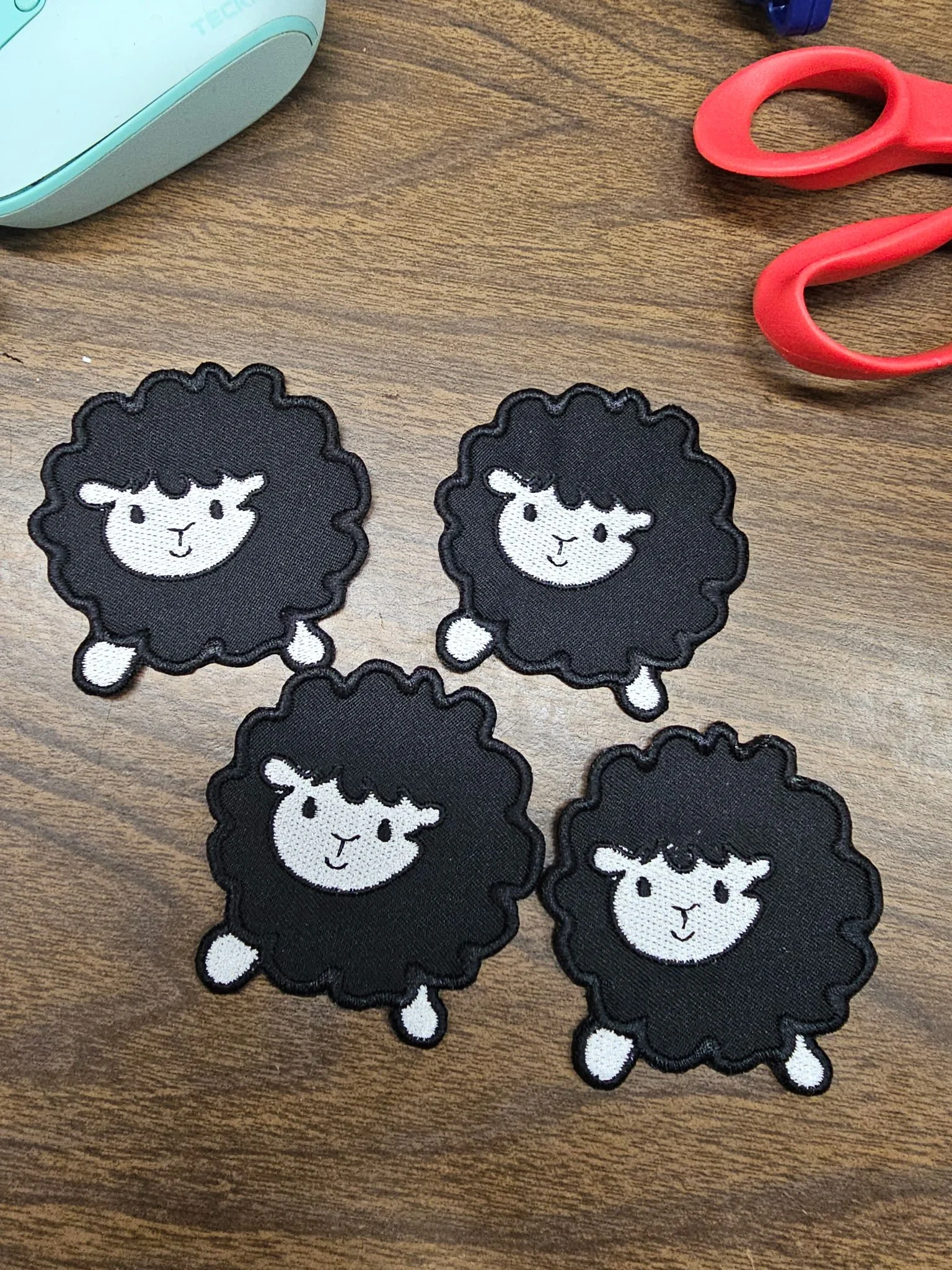Four black embroidered patches of sheep with white faces and legs on a wooden table, with a pair of red scissors and part of a teal item visible.