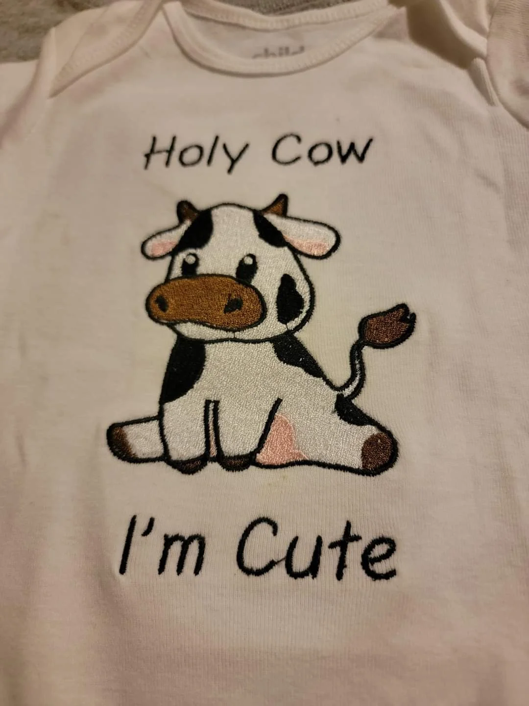 A white baby shirt with a cute cartoon cow illustration and the text "Holy Cow I'm Cute."