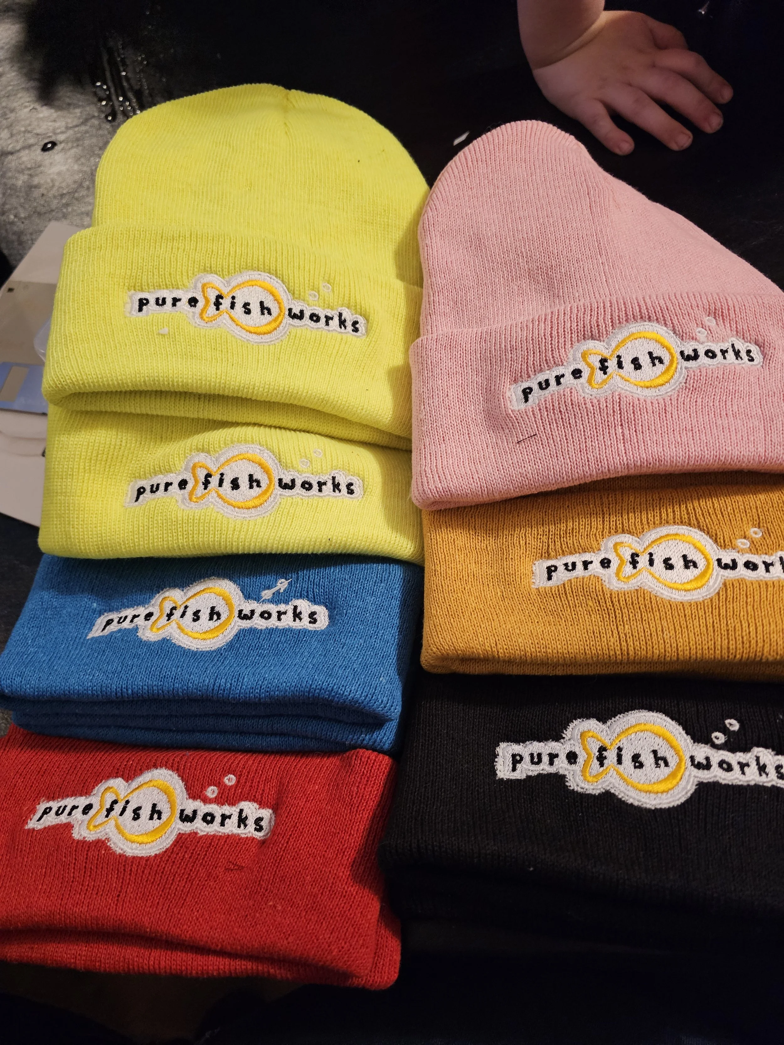 Stack of colorful beanies with 'Pure Fish Works' logo on each, in yellow, pink, blue, red, orange, and black.