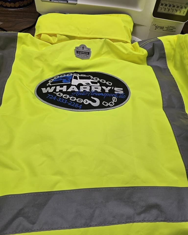 High-visibility yellow safety vest with black and blue embroidered patch of a tow truck and the words "Wharry's CEG e9, LLC" along with a phone number, placed on a table near a sewing machine.