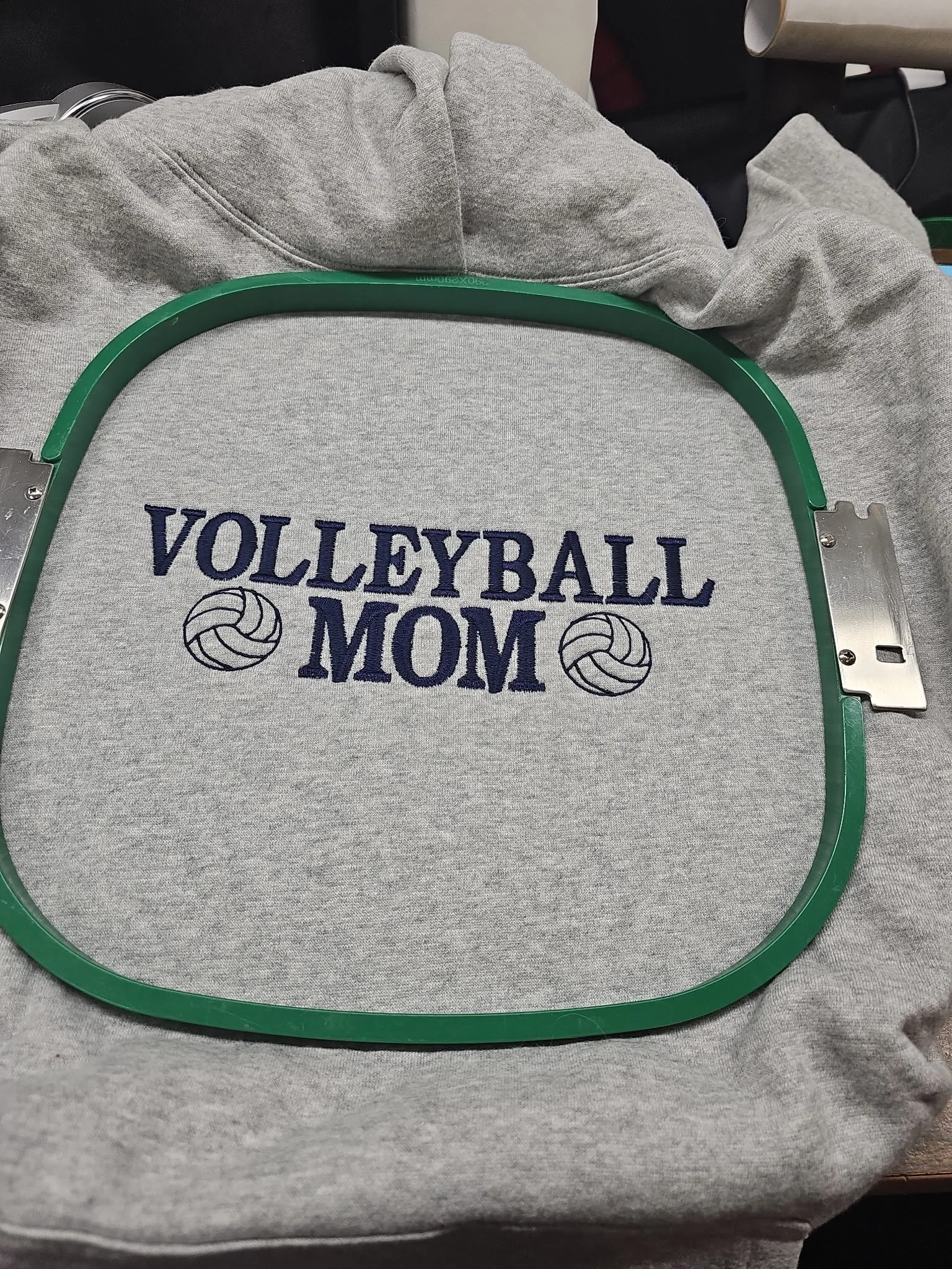Gray sweatshirt with embroidery that reads 'VOLLEYBALL MOM' with two volleyball icons on either side of the word 'MOM', and an embroidery hoop placed over it.