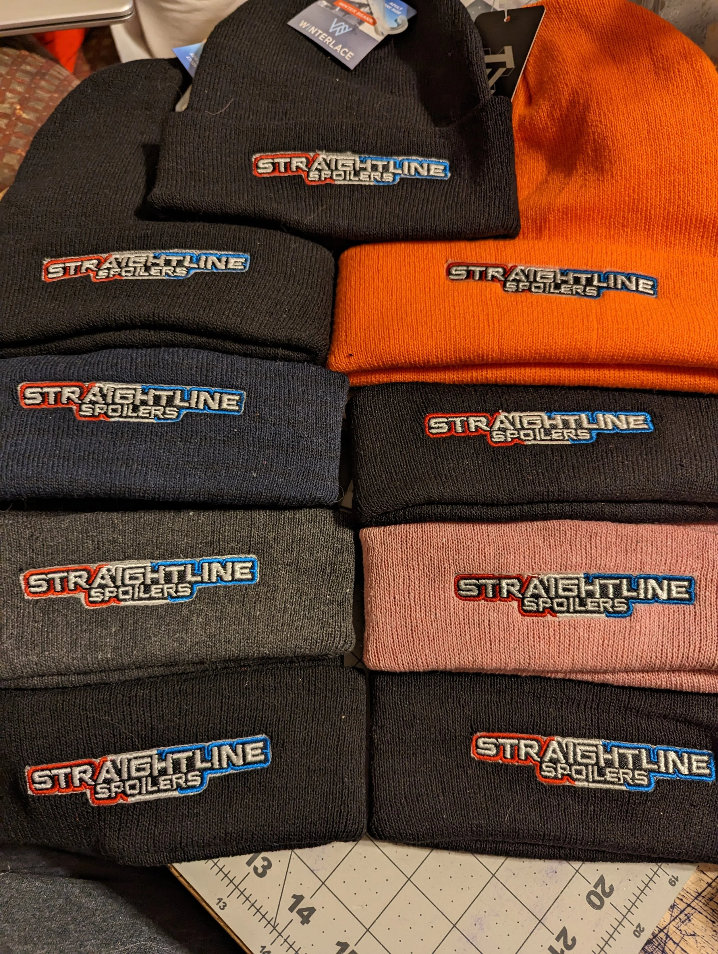 A display of nine beanies with embroidered logos reading 'Straightline Spoilers,' arranged in three rows. The beanies are in black, dark gray, orange, pink, and other colors.