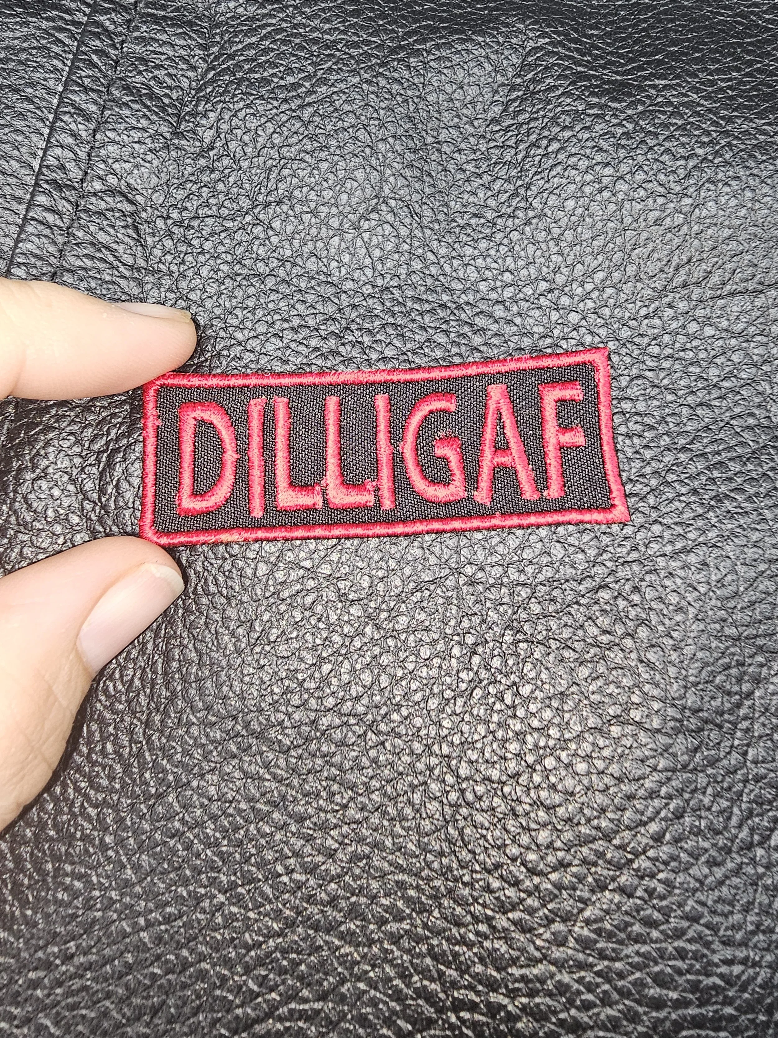 Close-up of a black leather surface with a pink embroidered patch that reads 'DILLGAF'.