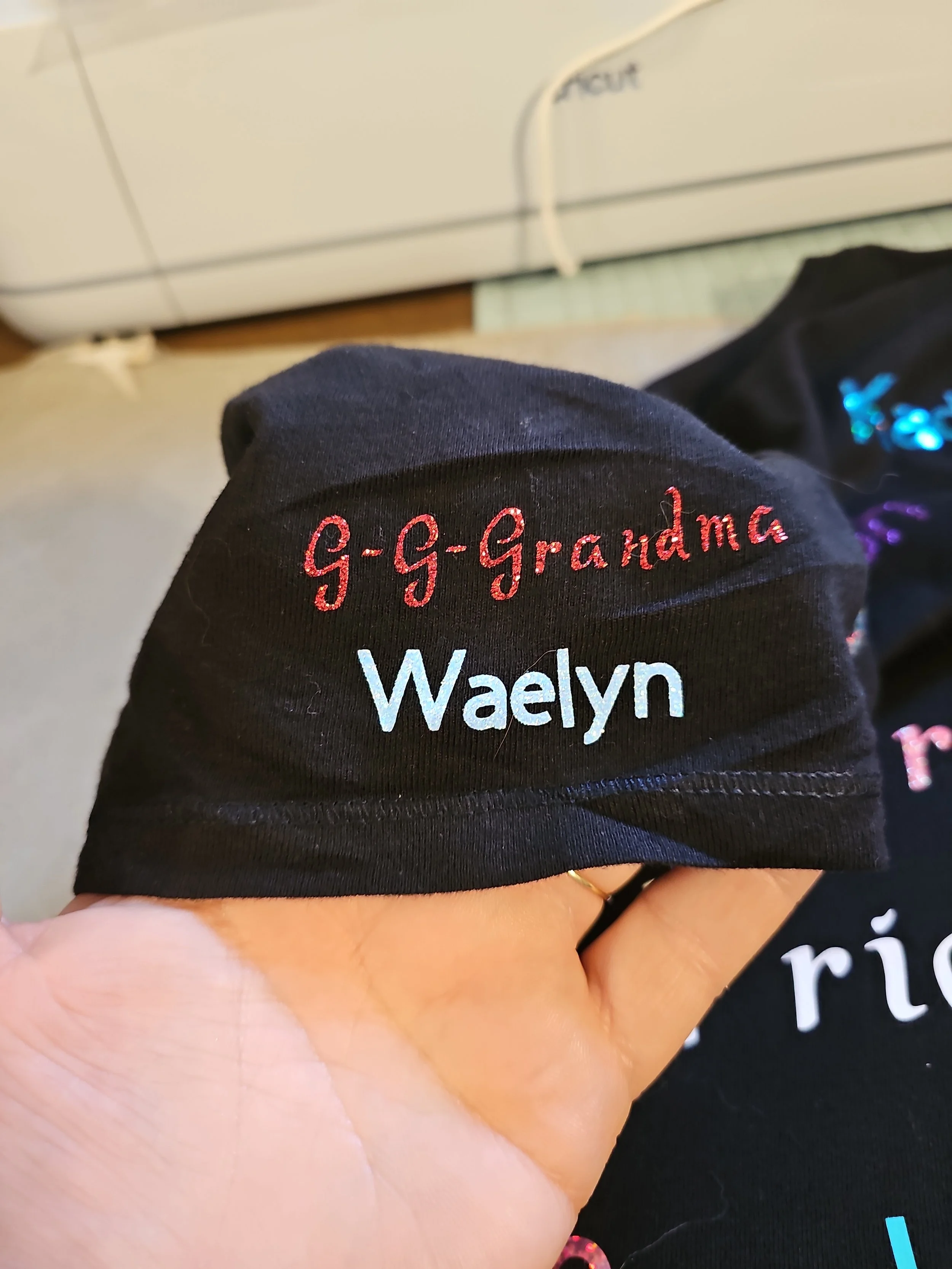 Black fabric with embroidered text in pink and white reading 'G-G-Grandma' and 'Waelyn,' held in a person's hand.