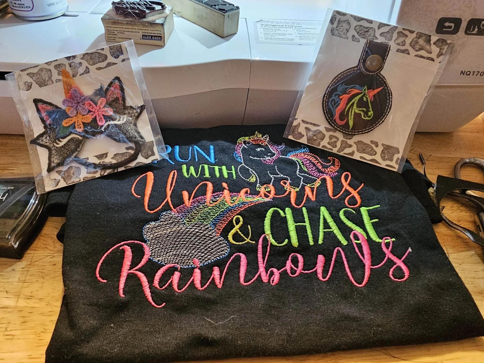 Embroidered black tote bag with colorful rainbow, clouds, unicorns, and the words "Run with Unicorns & Chase Rainbows." Two embroidered patches: one unicorn head with colorful hair, and one star-shaped patch with rainbow colors and flowers, in packag