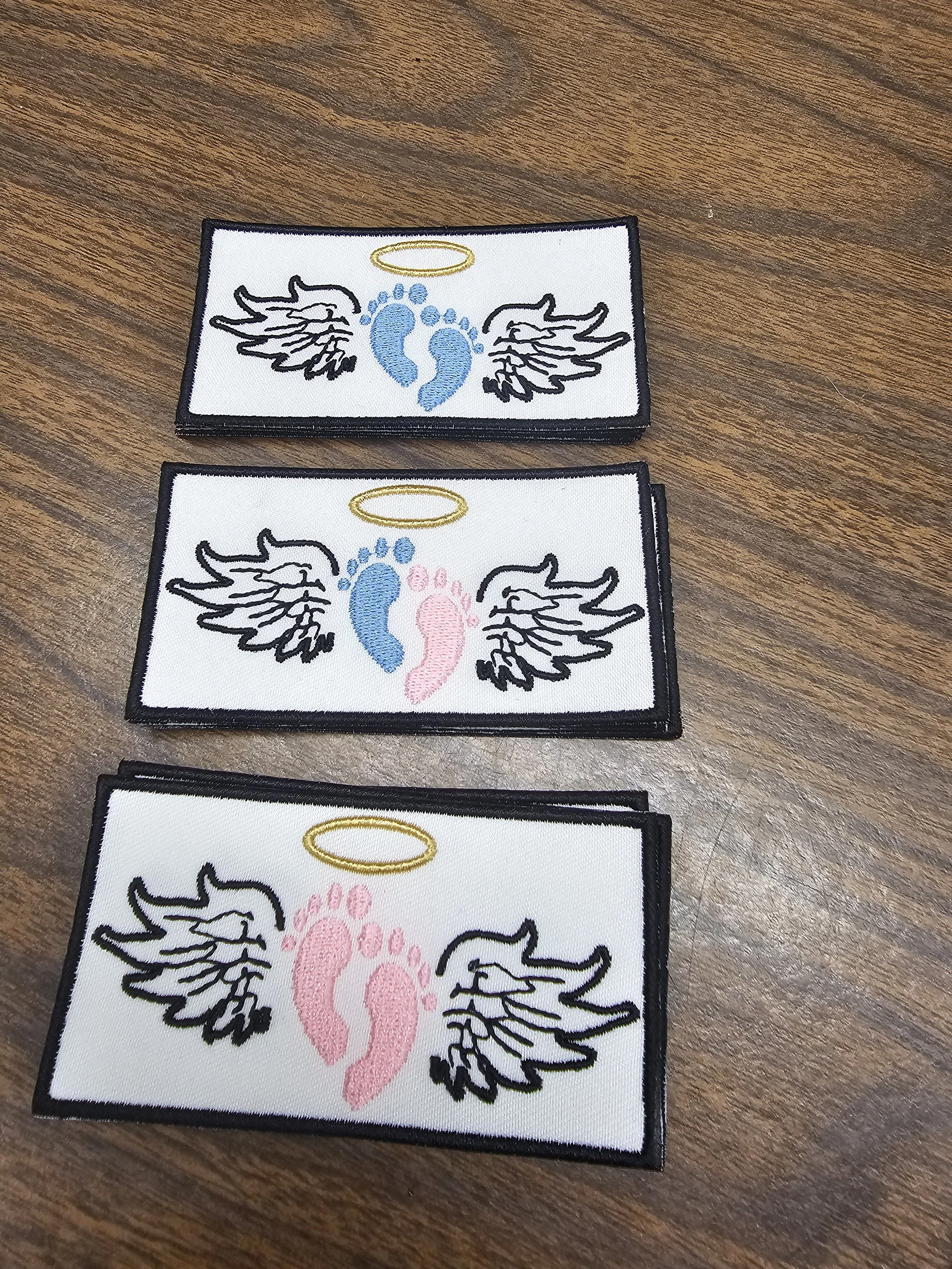 Three embroidered patches with angelic wing and halo designs, each featuring different colored footprints - blue, pink, and a darker pink, on a wooden surface.