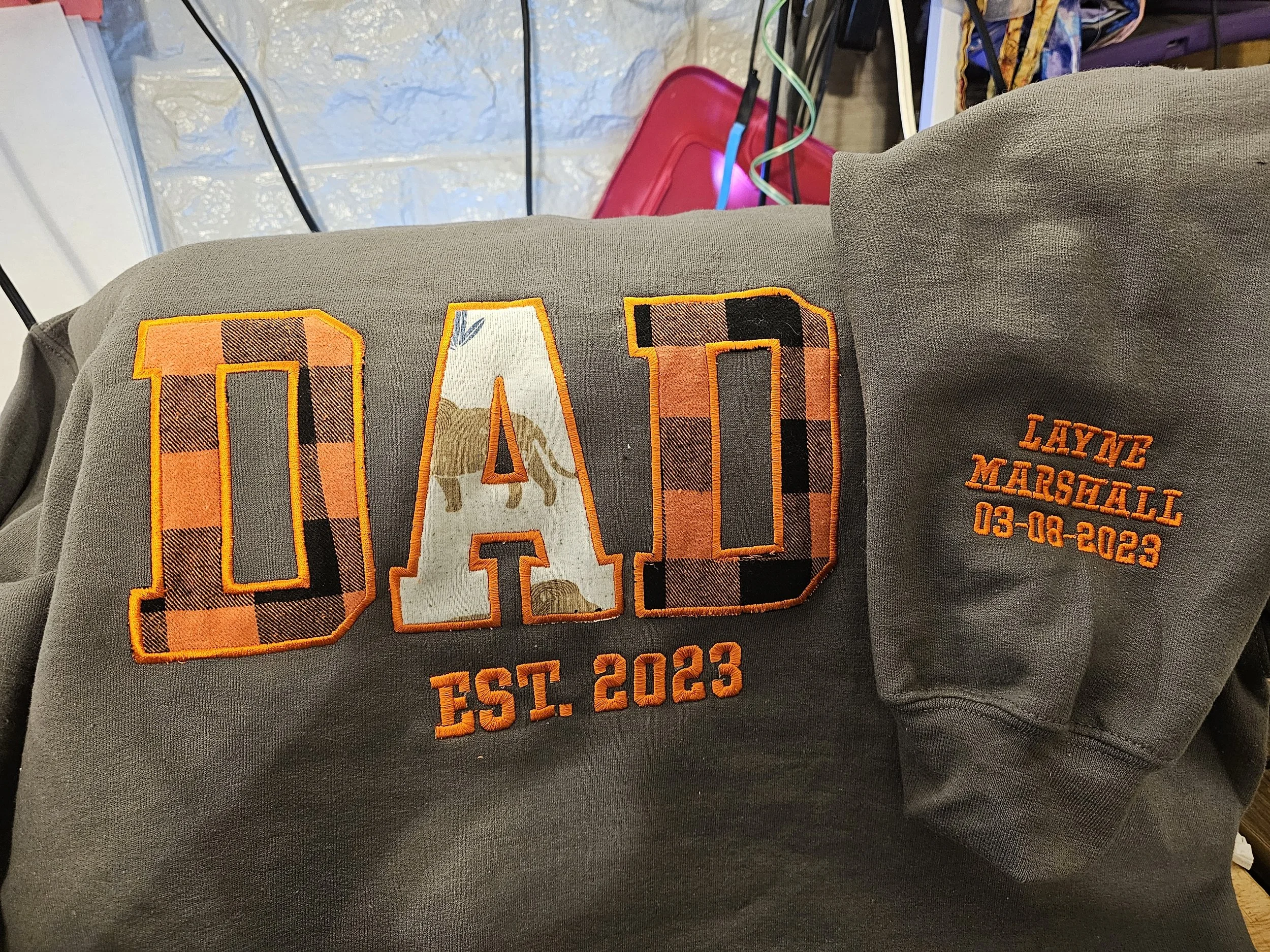 Close-up of a dark gray sweatshirt with orange embroidered text and patches. The large front patch reads 'DAD' with buffalo plaid pattern and a lion graphic inside the letter 'A'. To the right, smaller orange embroidered text states 'Laynce Marshall