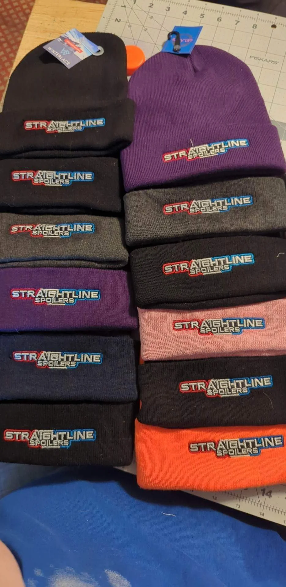 A collection of beanies with 'Straightline Spoilers' embroidered on the front, arranged in two columns on a work surface.