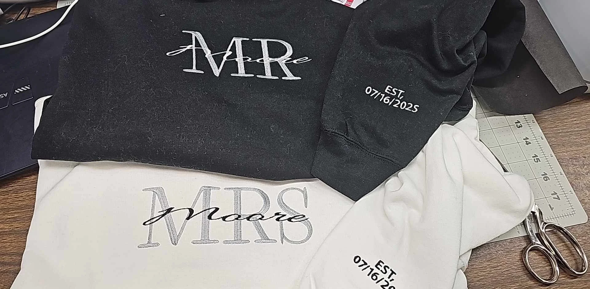 Two fabric items with embroidery; one black, one white. The black fabric has the embroidered words 'AARre' and 'EST 07/16/2025.' The white fabric has the embroidered words 'make' and a partially visible date. A pair of scissors is placed next to the