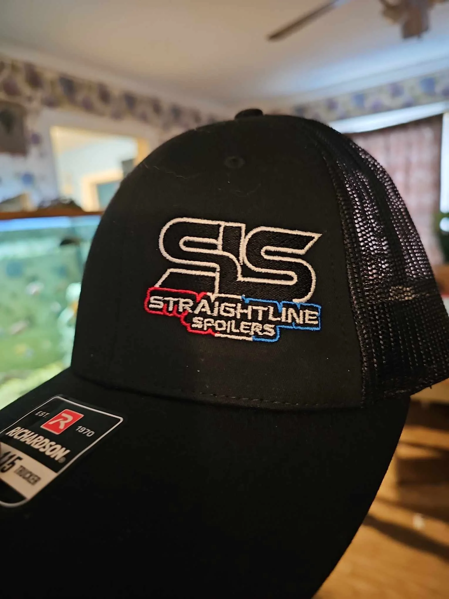 Black trucker cap with embroidered logo reading 'SS Straightline Spoilers' in white, red, and blue thread, front view, indoors with background including a fish tank and a window.