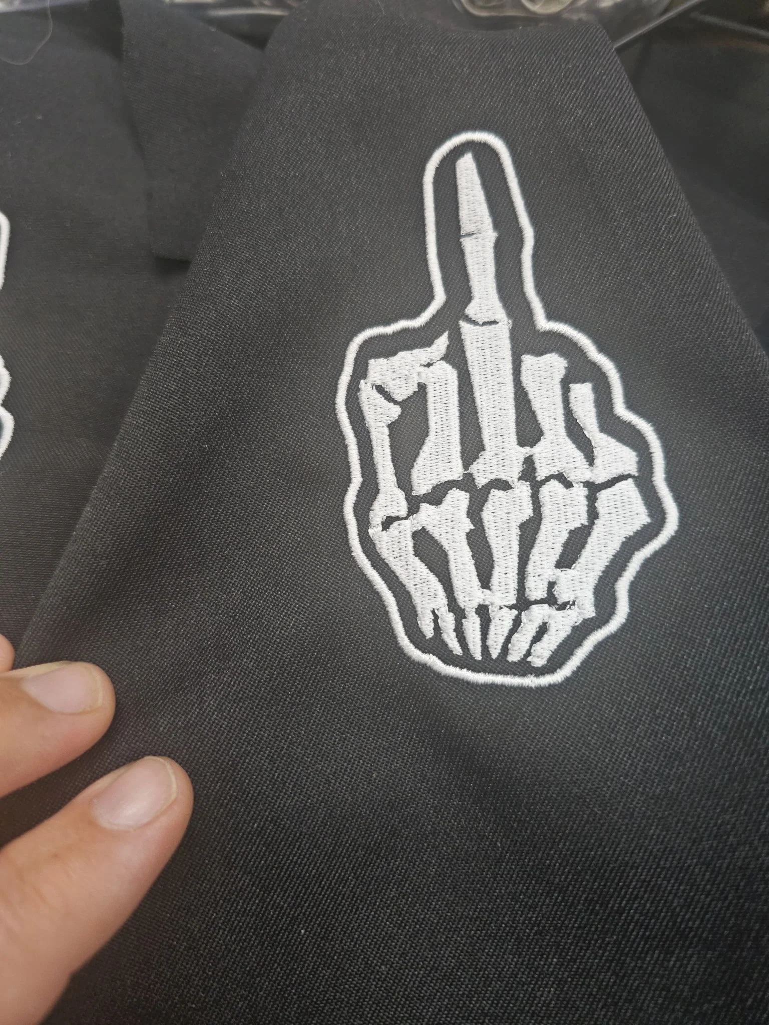 Black fabric with a white embroidered design of a skeleton hand making an obscene gesture with the middle finger extended.