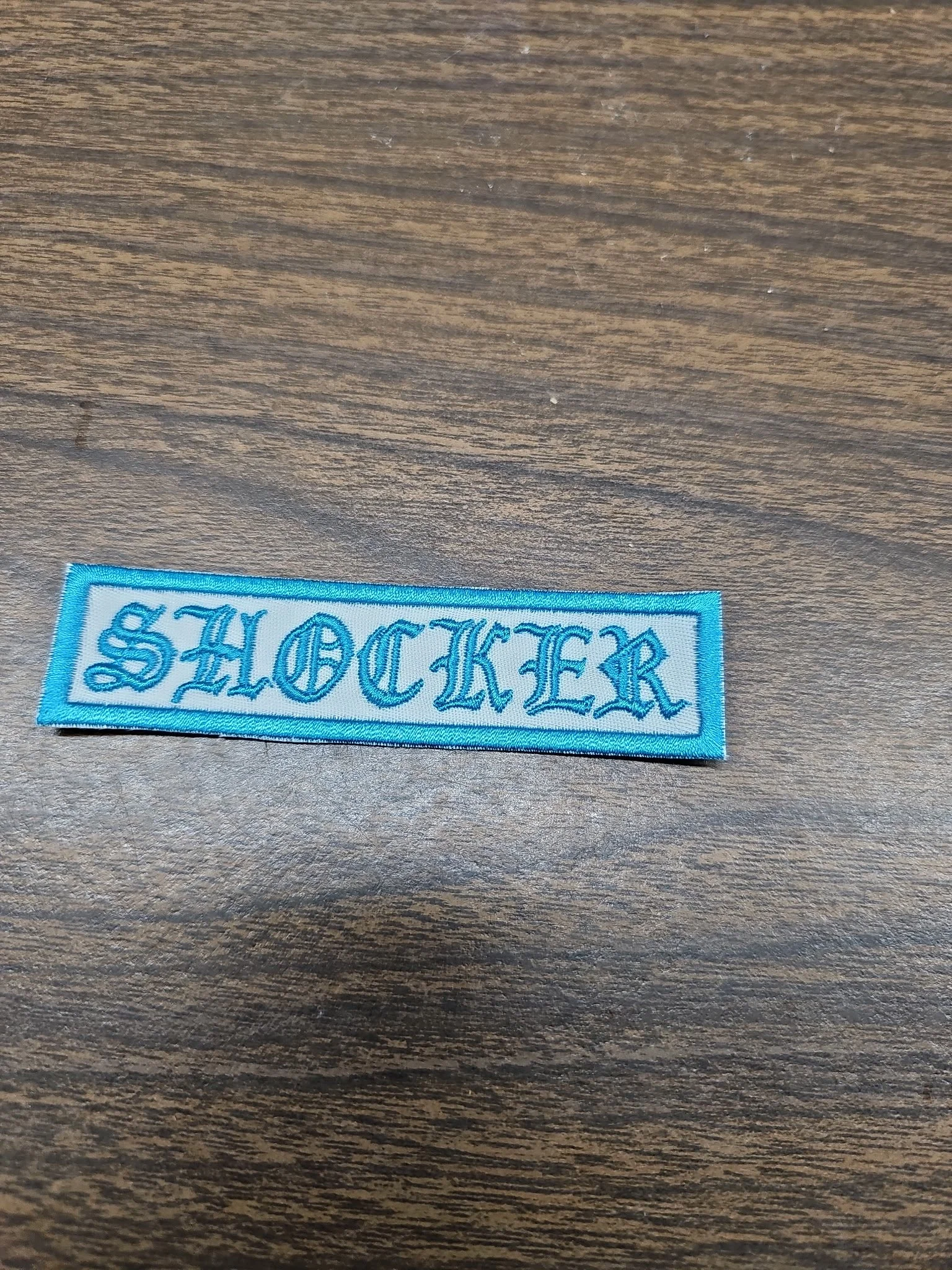 A light blue embroidered patch with the word 'SHOCKER' in old English font, placed on a wooden surface.