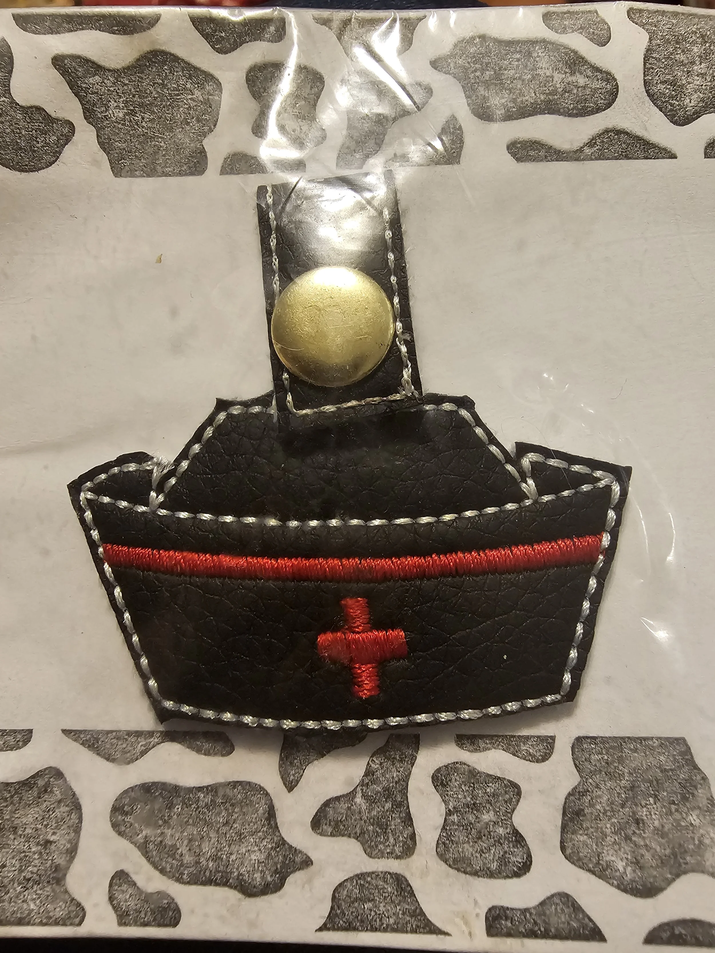 A small embroidered patch of a black nurse's cap with red cross and red line, attached to a plastic backing with a metallic snap button.