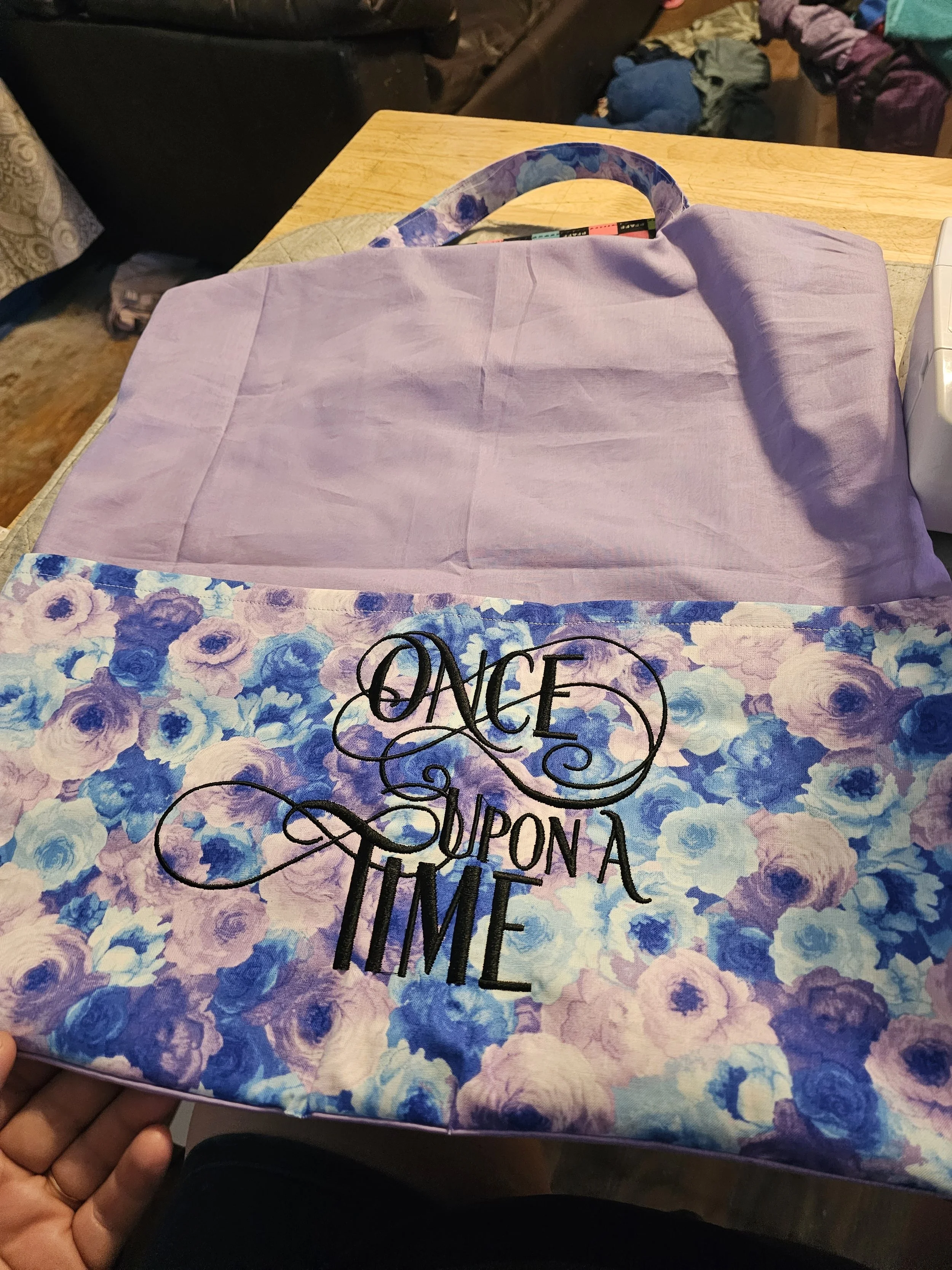 A tote bag with a purple floral pattern and the phrase 'Once Upon a Time' printed in black, held over a pink fabric surface on a wooden table.