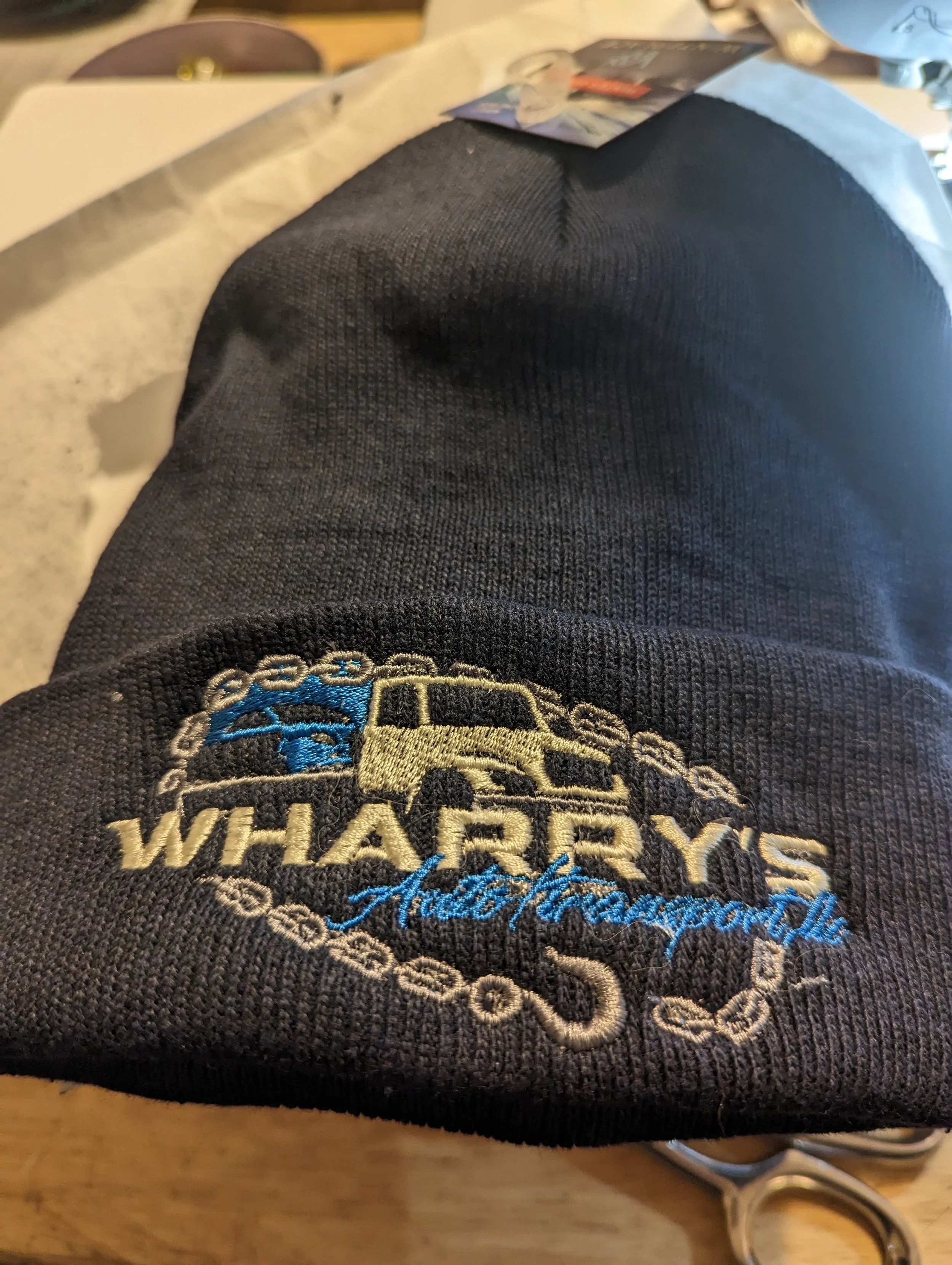 Close-up of a black baseball cap with embroidered logo reading "WHARREYS Auto Racing" featuring a yellow car, with a chain design surrounding the text, placed on a surface with some items in the background.