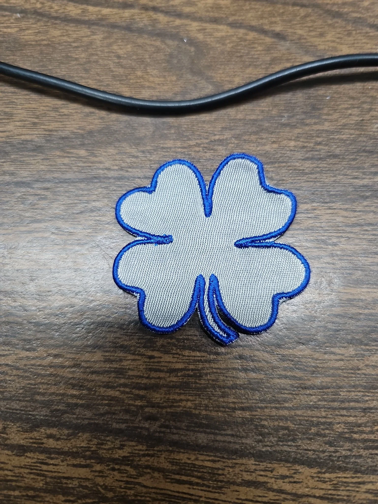 A white embroidered patch in the shape of a four-leaf clover with blue outline, placed on a wooden surface.