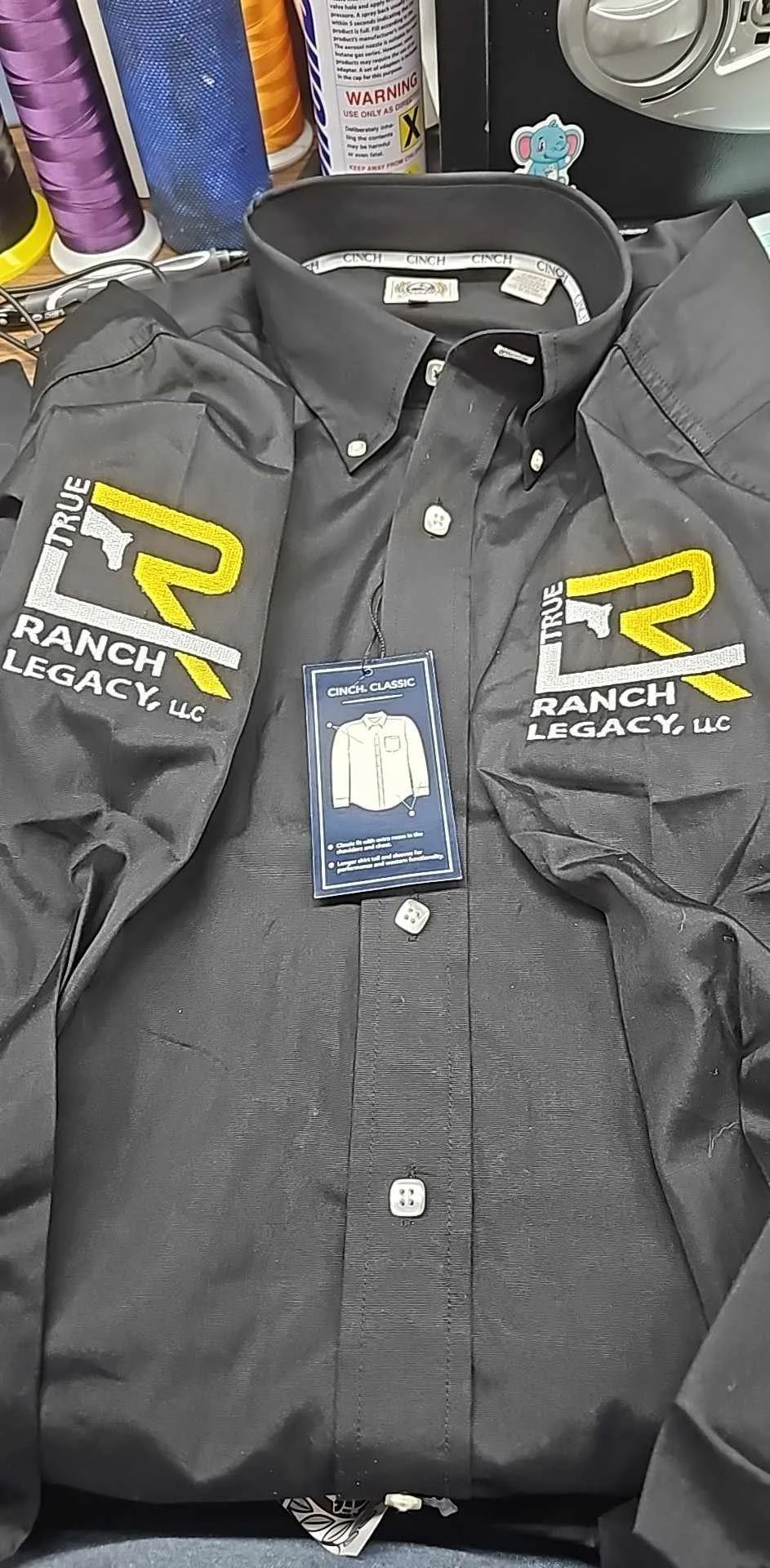 A black button-up shirt with embroidered patches on the sleeves reading 'True Ranch Legacy LLC' and a yellow 'R' logo, laying on a work surface with sewing supplies and tools around.