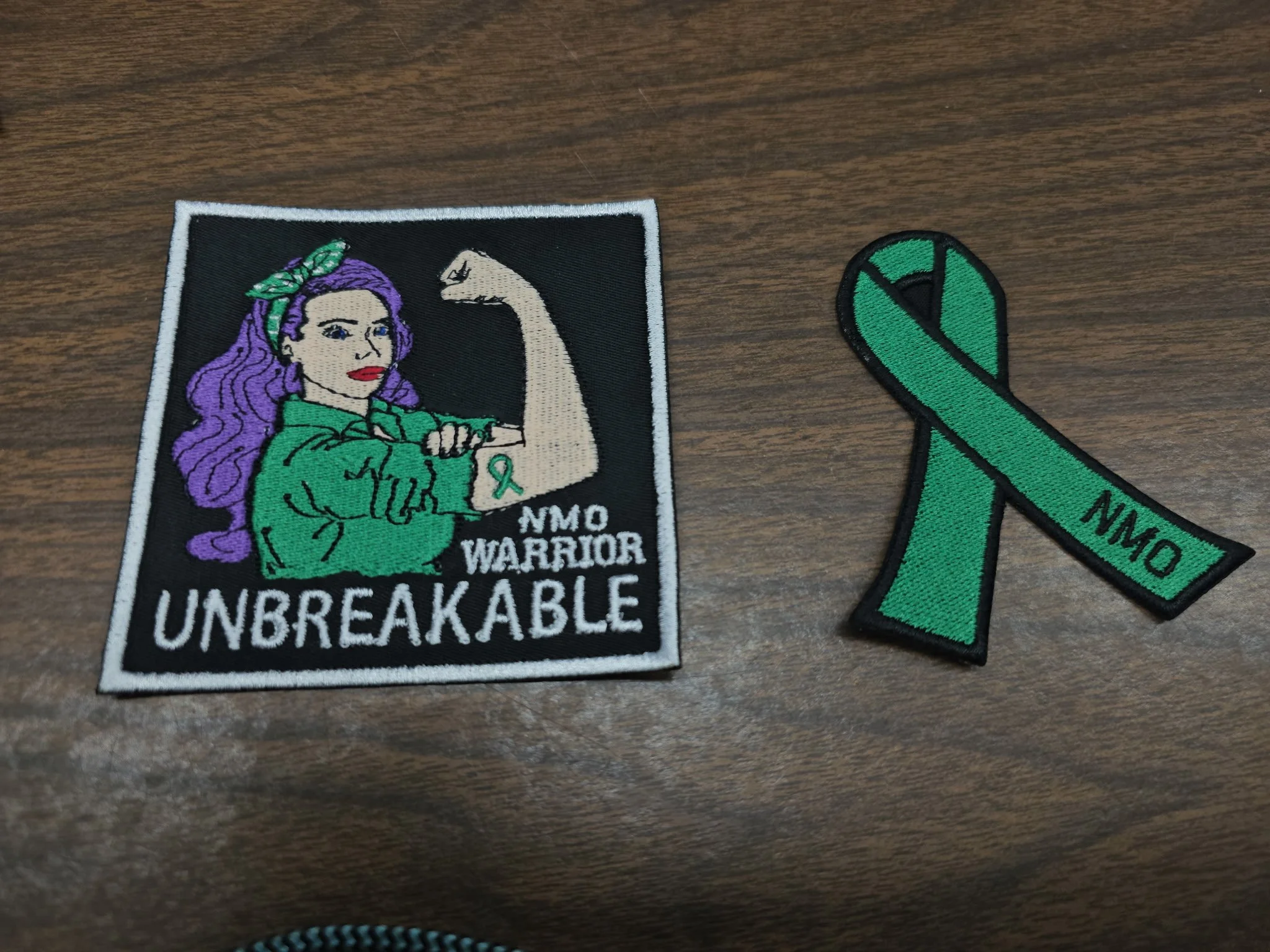 Two patches on a wooden surface. The one on the left depicts a woman with purple hair in a green shirt, flexing her arm with a white swagger and a small green ribbon, with text that reads "NMO Warrior Unbreakable." The patch on the right is a green a
