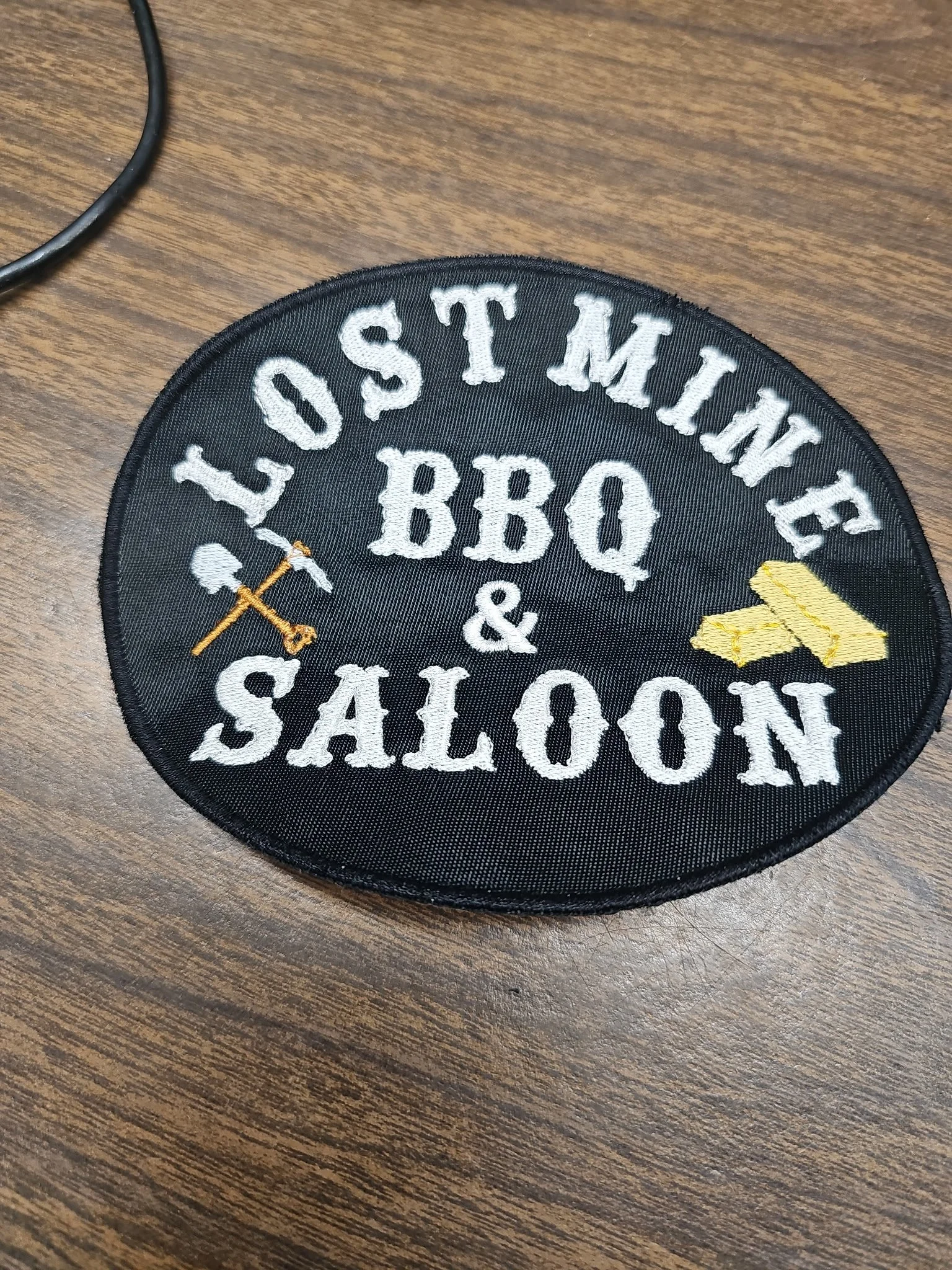 A round embroidered patch with a black background, white text that reads 'Lost Mine BBQ & Saloon', a small yellow paper boat, and a crossed fork and sword design.