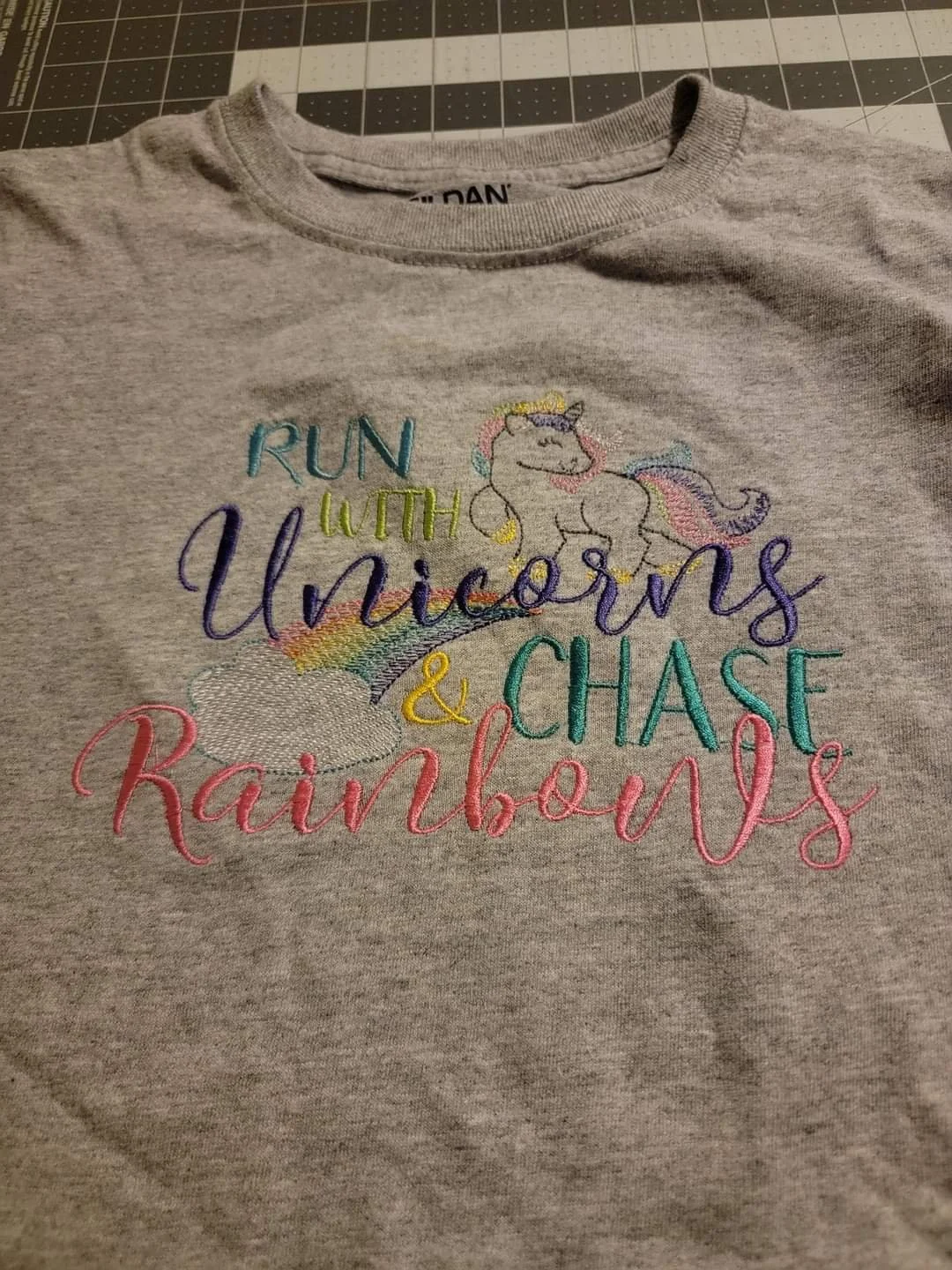 Gray T-shirt with colorful embroidered design featuring a unicorn, a rainbow, clouds, and the phrase "Run with Unicorns & Chase Rainbows" in various bright colors.