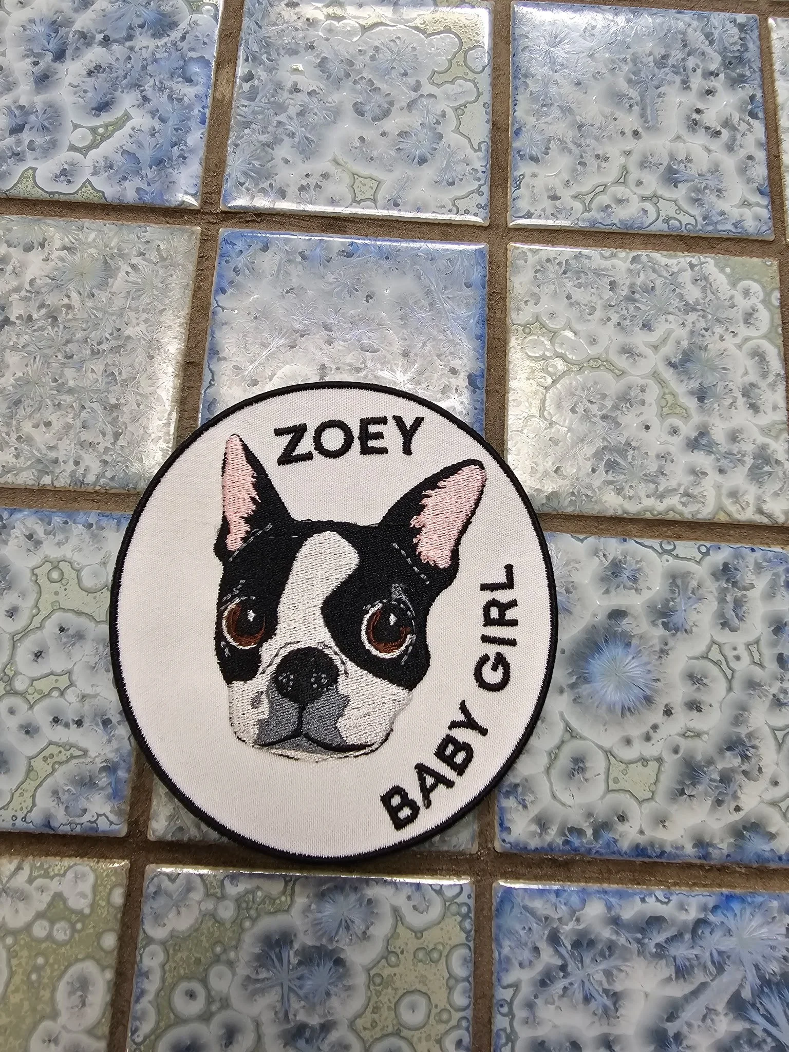 Patch with a Boston Terrier's face, black and white, and the text 'ZoeY Baby Girl' around the face, on a tiled surface with blue and white patterns.