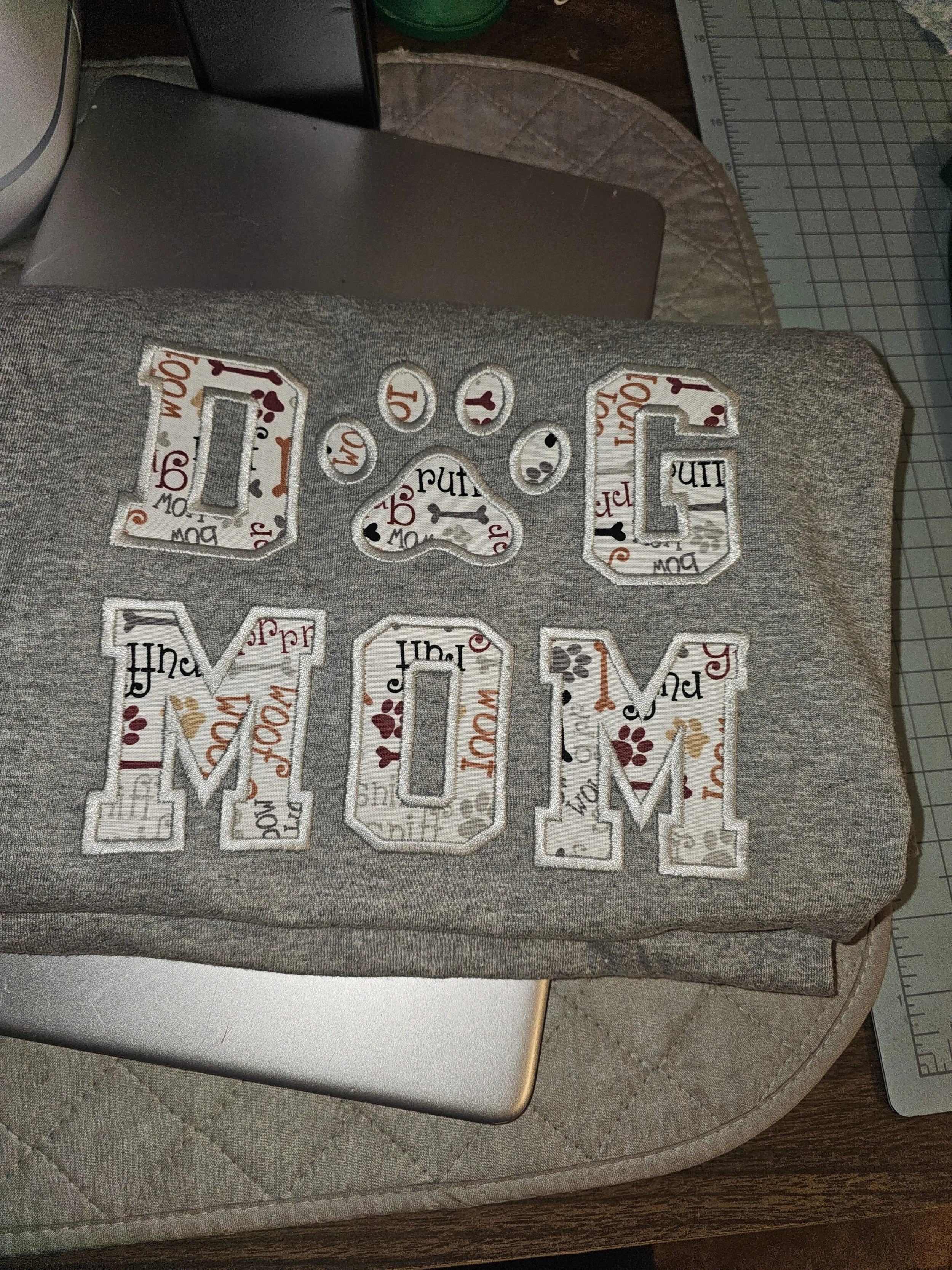 Gray fabric with embroidered letters spelling 'LOVE MOM' on top of a silver laptop, surrounded by crafting tools.