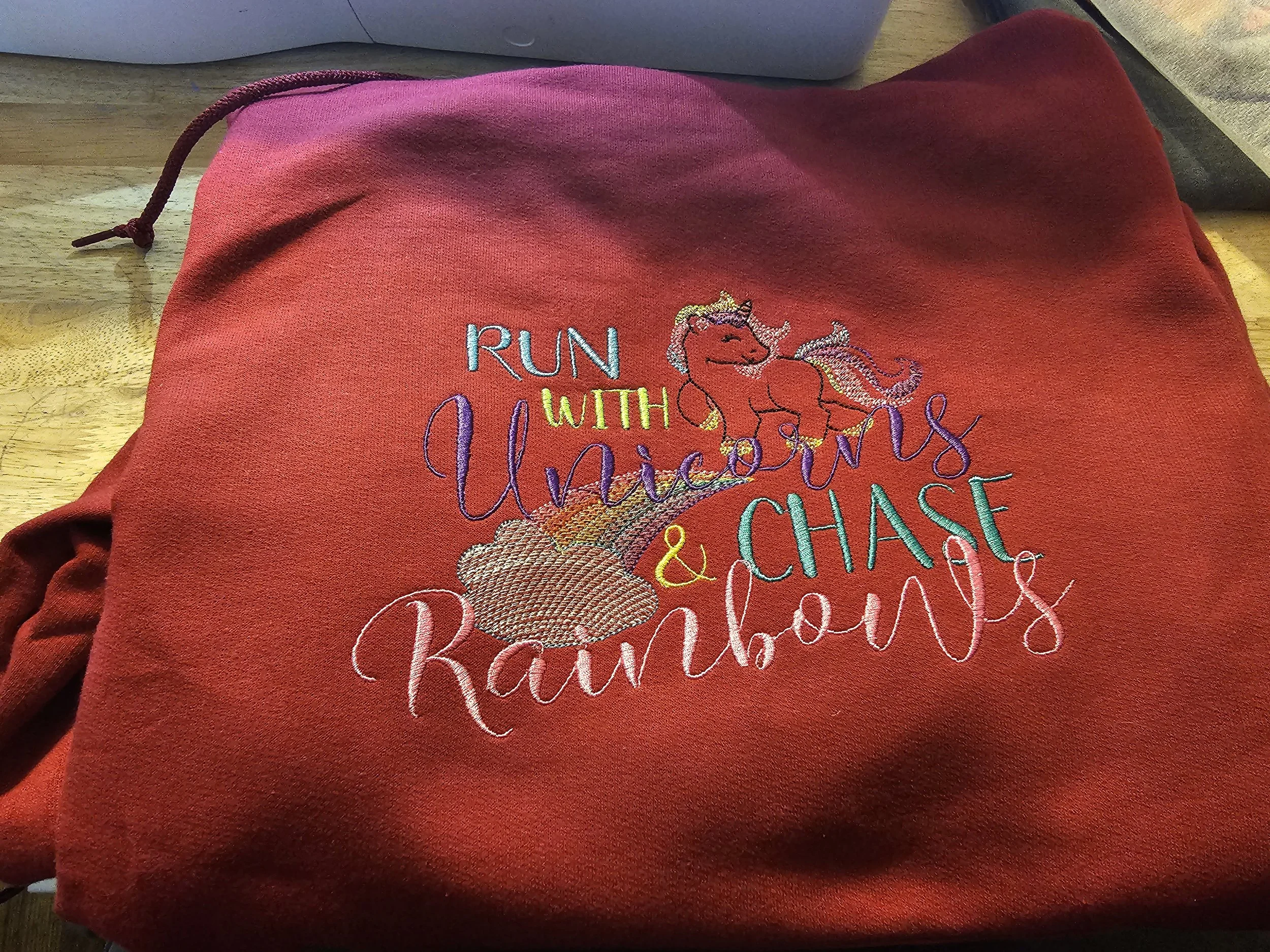 Red hoodie with embroidered design of a unicorn, rainbow, and clouds, and the words "Run with Unicorns & Rainbow".