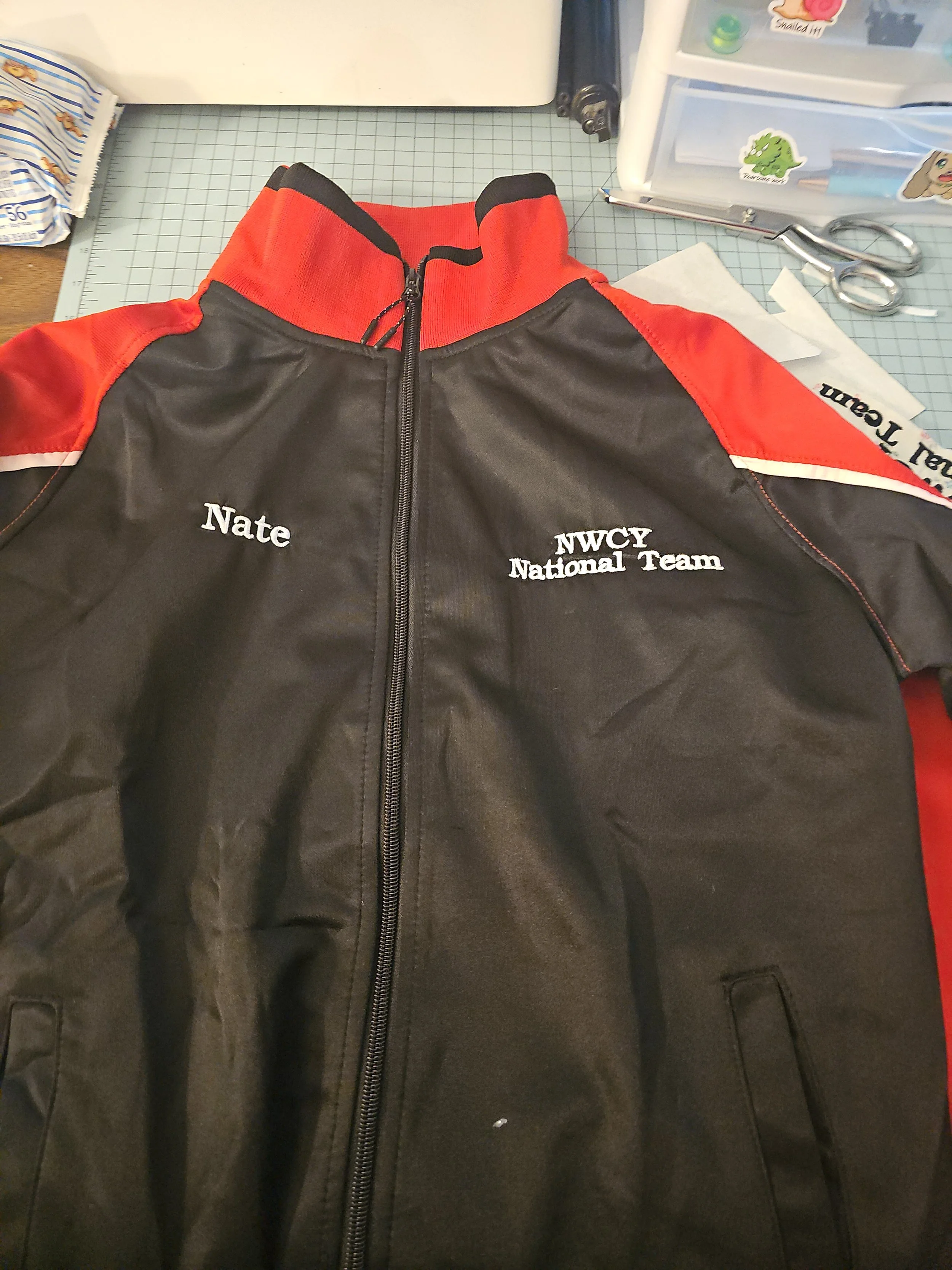 Black and red jacket with embroidered text 'Nate' on the left chest and 'NWCGY National Team' on the right chest, laid flat on a work surface with scissors, paper, and a plastic storage container around.