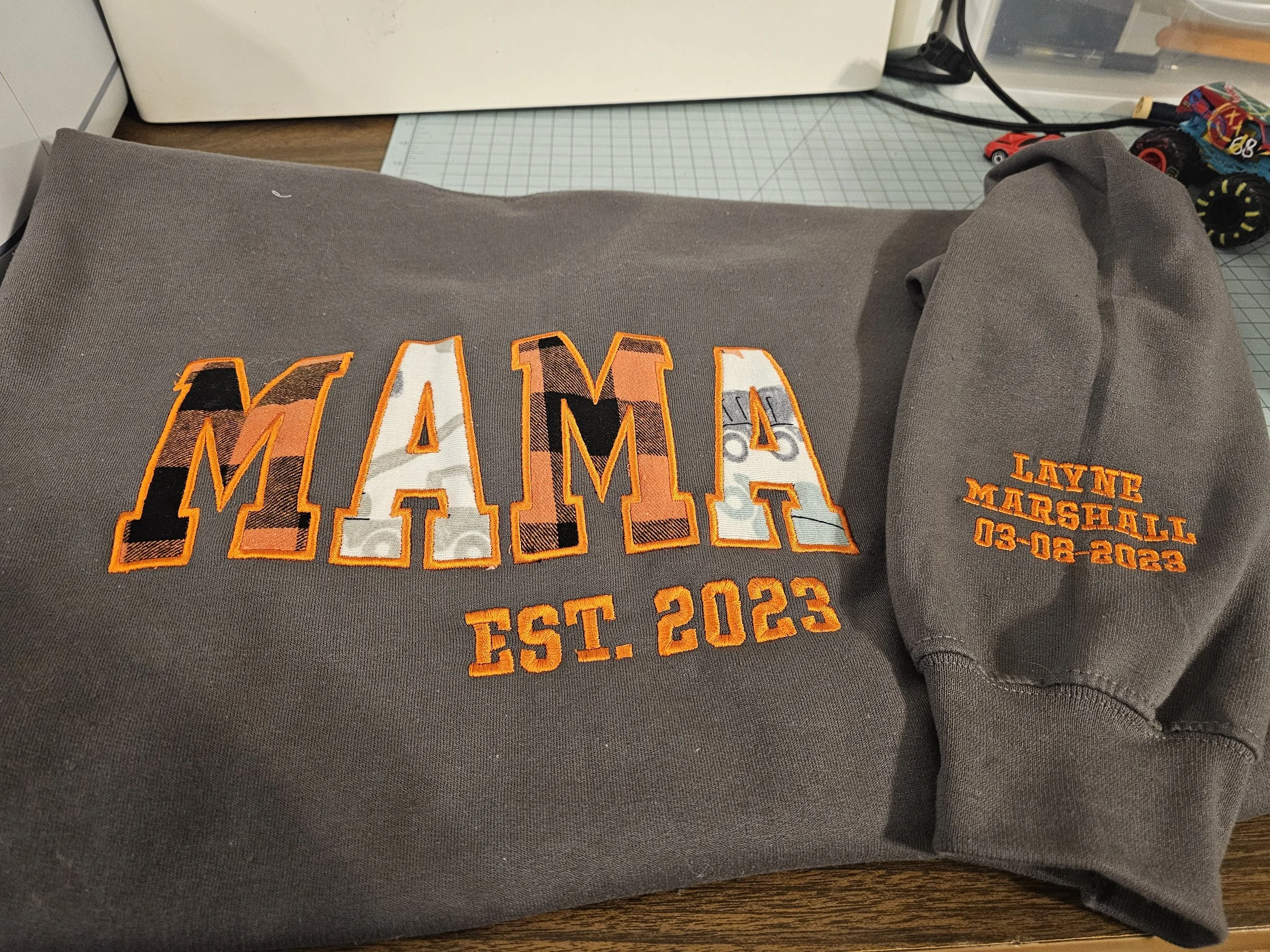 Gray hoodie with 'MAMA' embroidered in large multicolored letters and 'EST. 2023' below, along with a matching gray hat with orange embroidered text 'LAVNE MARSHALL 03-08-2023' on the side.