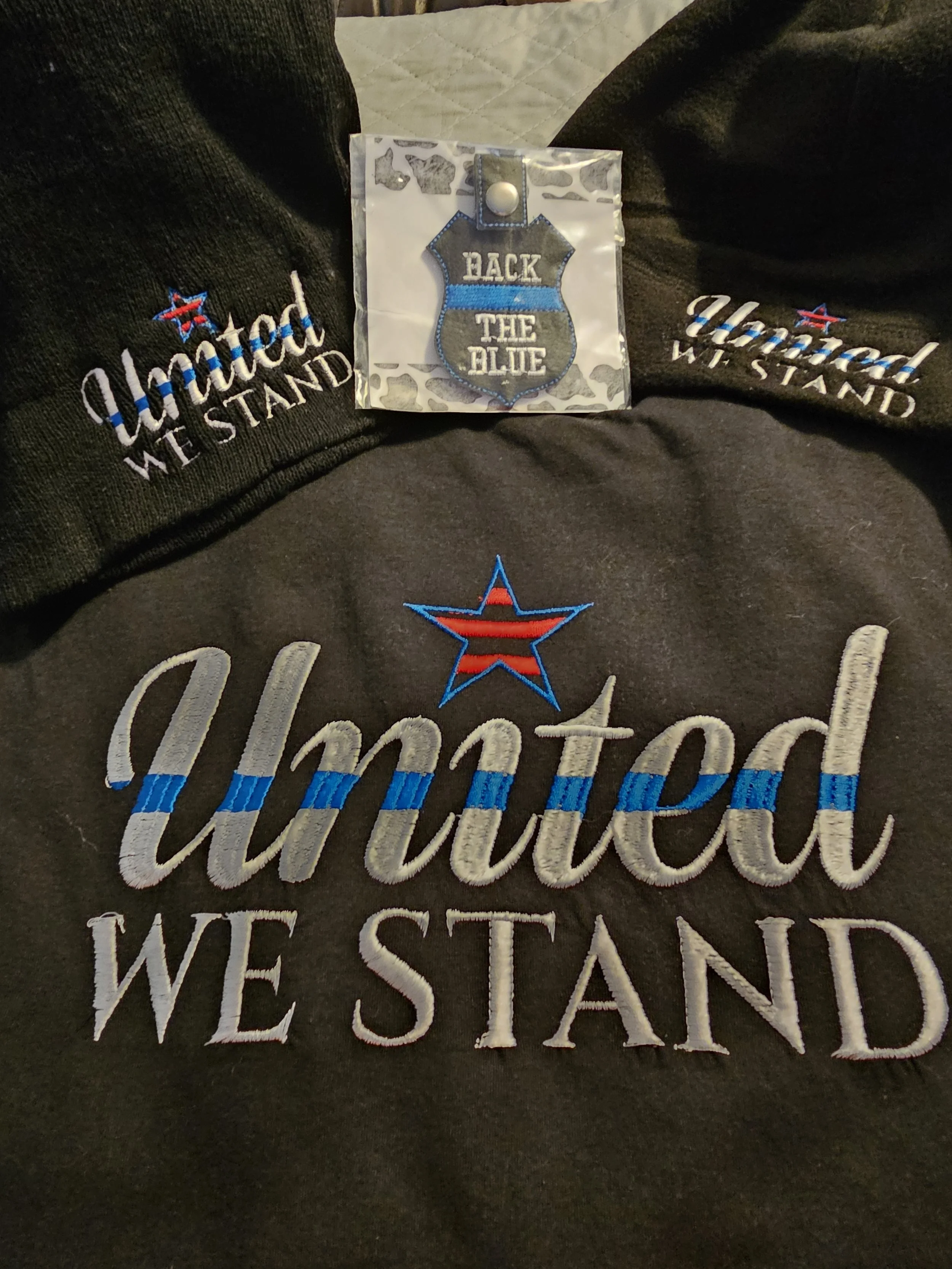 A black sweatshirt with embroidered text that reads 'United We Stand' and an embroidered star. A small badge with blue and silver colors that reads 'Back the Blue' is placed on the sweatshirt, and two black hats embroidered with the same 'United We S