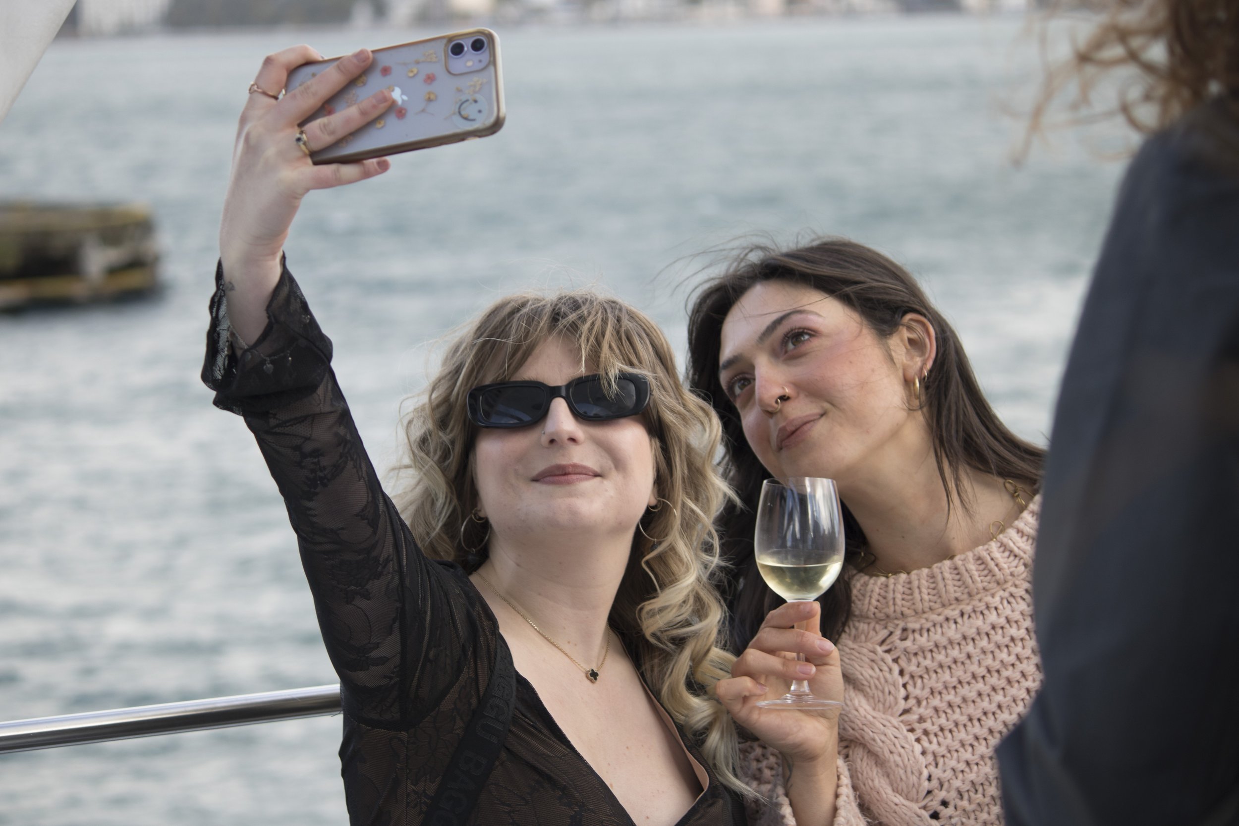 Two women taking a selfie by the water, one with sunglasses and the other holding a glass of white wine.
