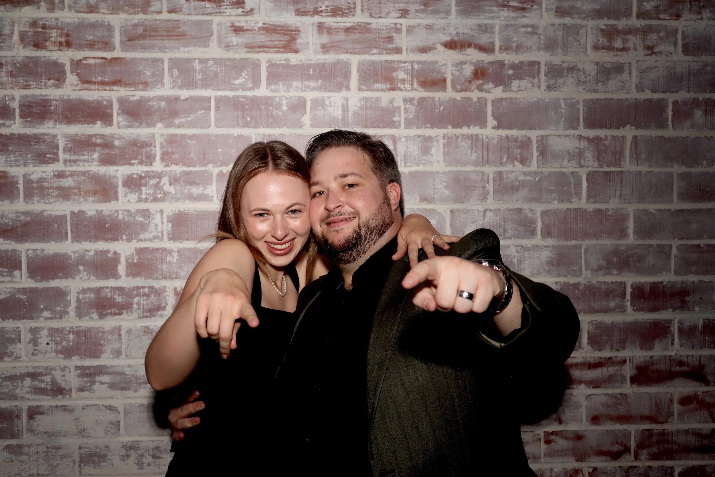A smiling woman and a man with a beard are posing together, pointing towards the camera, in front of a brick wall background.