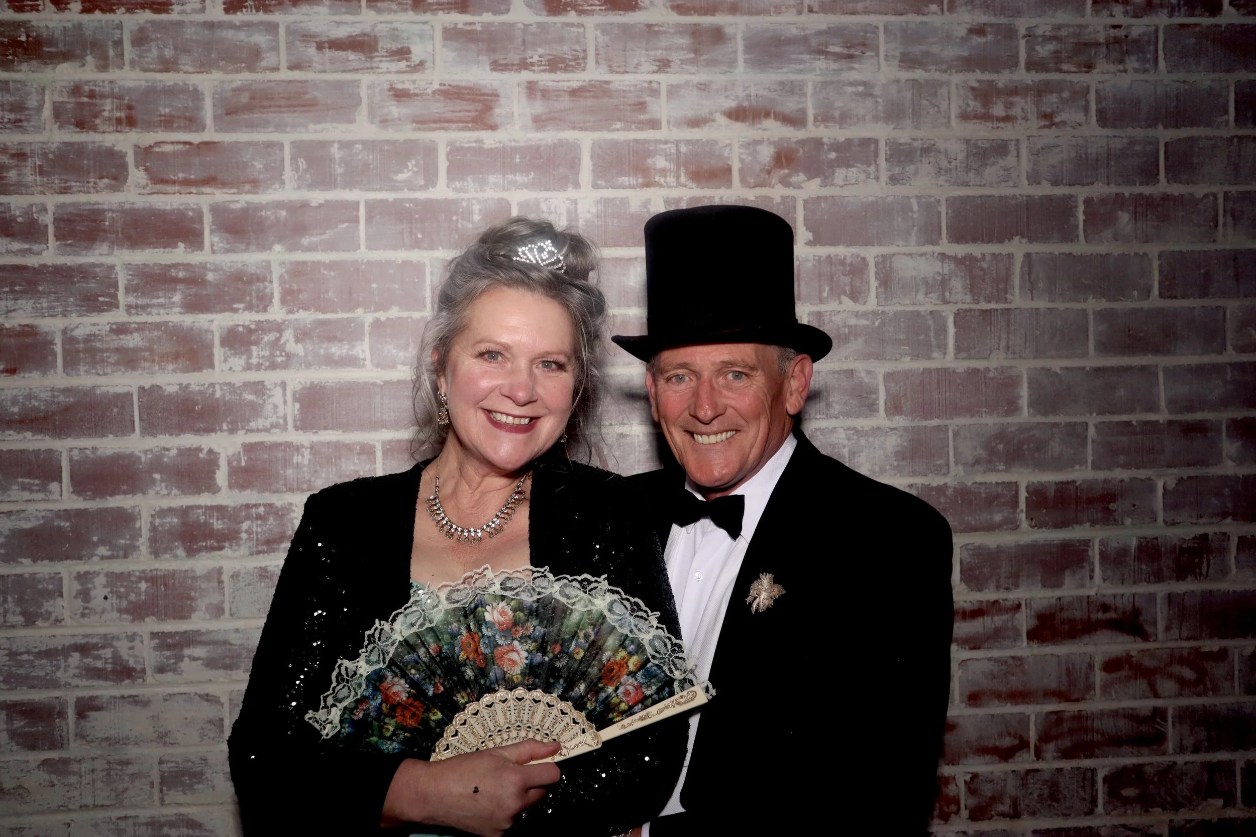 An older couple dressed in formal attire, smiling at the camera. The woman wears a tiara, a sparkling necklace, and a black dress with sequins, holding a decorative hand fan. The man wears a black top hat, tuxedo with a bow tie, and a star-shaped pin