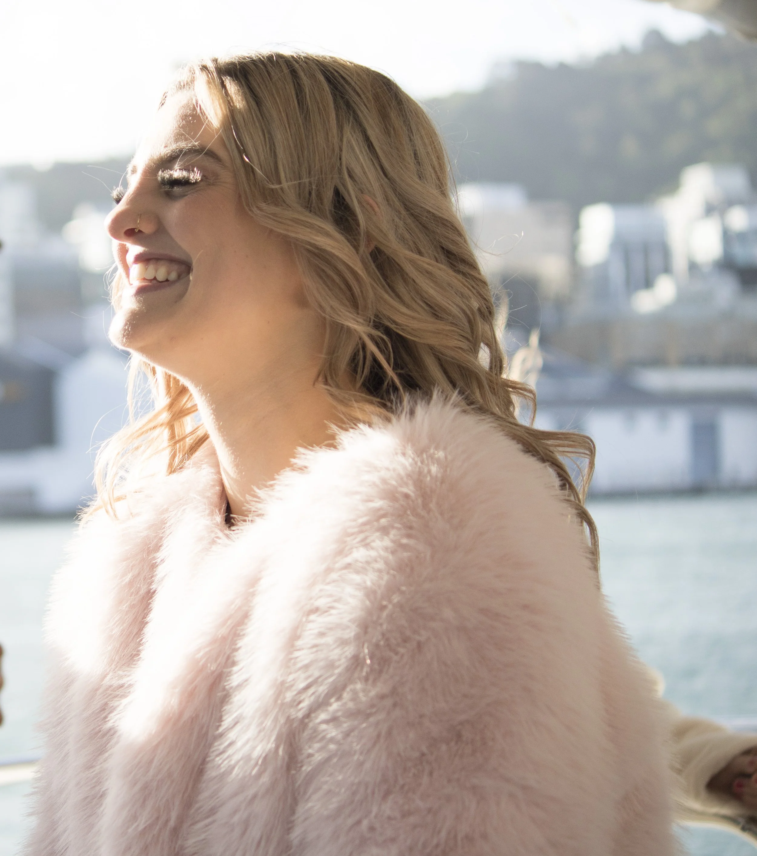 A smiling woman with wavy blonde hair wearing a pink fur coat with a city harbor in the background.