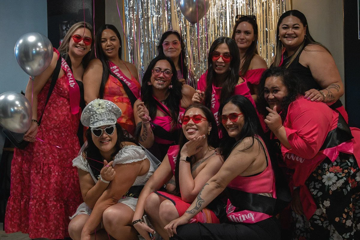 A group of women celebrating a bachelorette party, wearing pink and floral outfits, with some wearing red sunglasses and sash accessories, posing together indoors with balloons and metallic gold decorations in the background.
