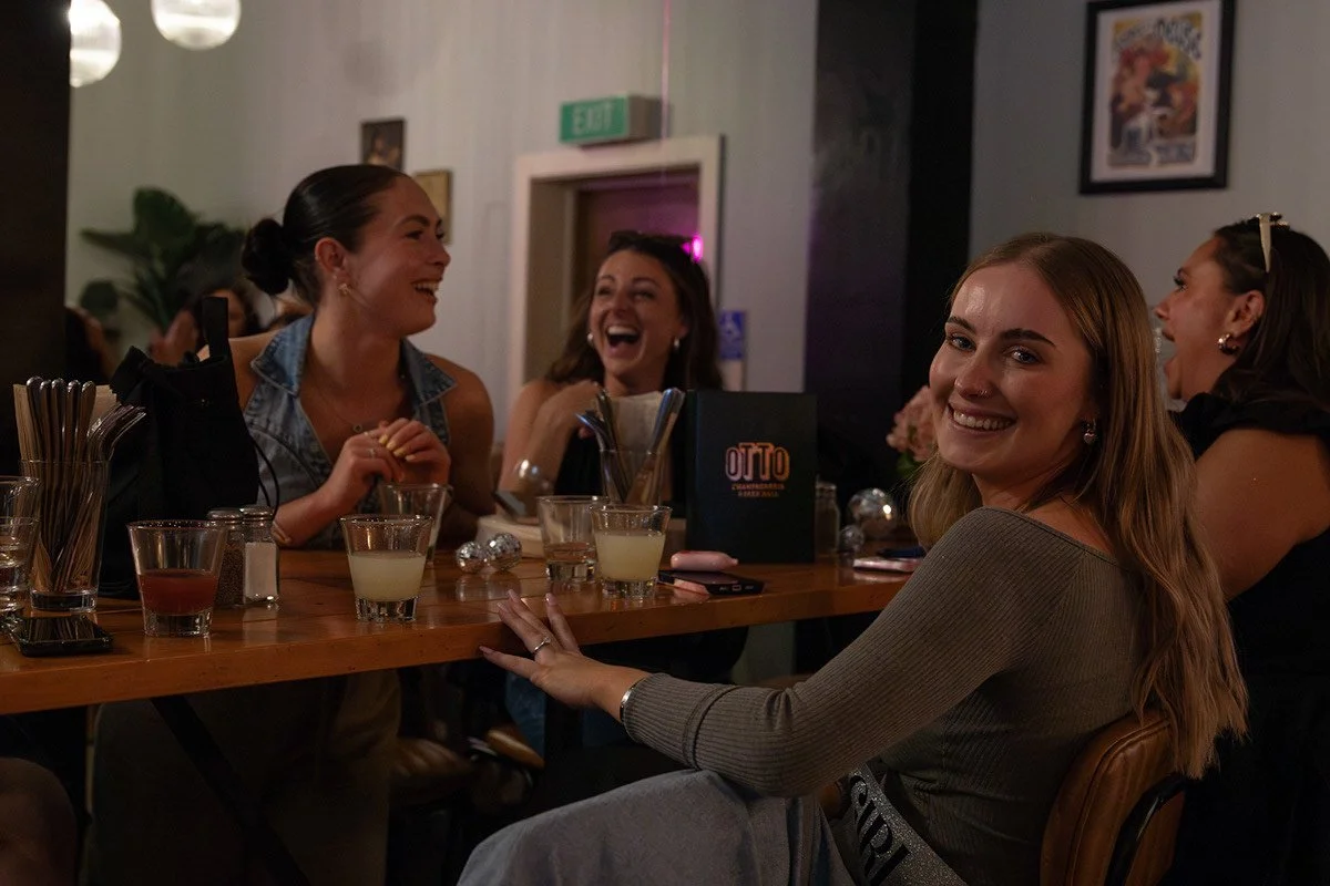 Four women sitting at a bar, smiling and laughing. The woman closest to the camera has long blonde hair, is wearing a gray top, and is touching the bar. The other three women have dark hair, are wearing sleeveless tops, and are engaged in conversatio