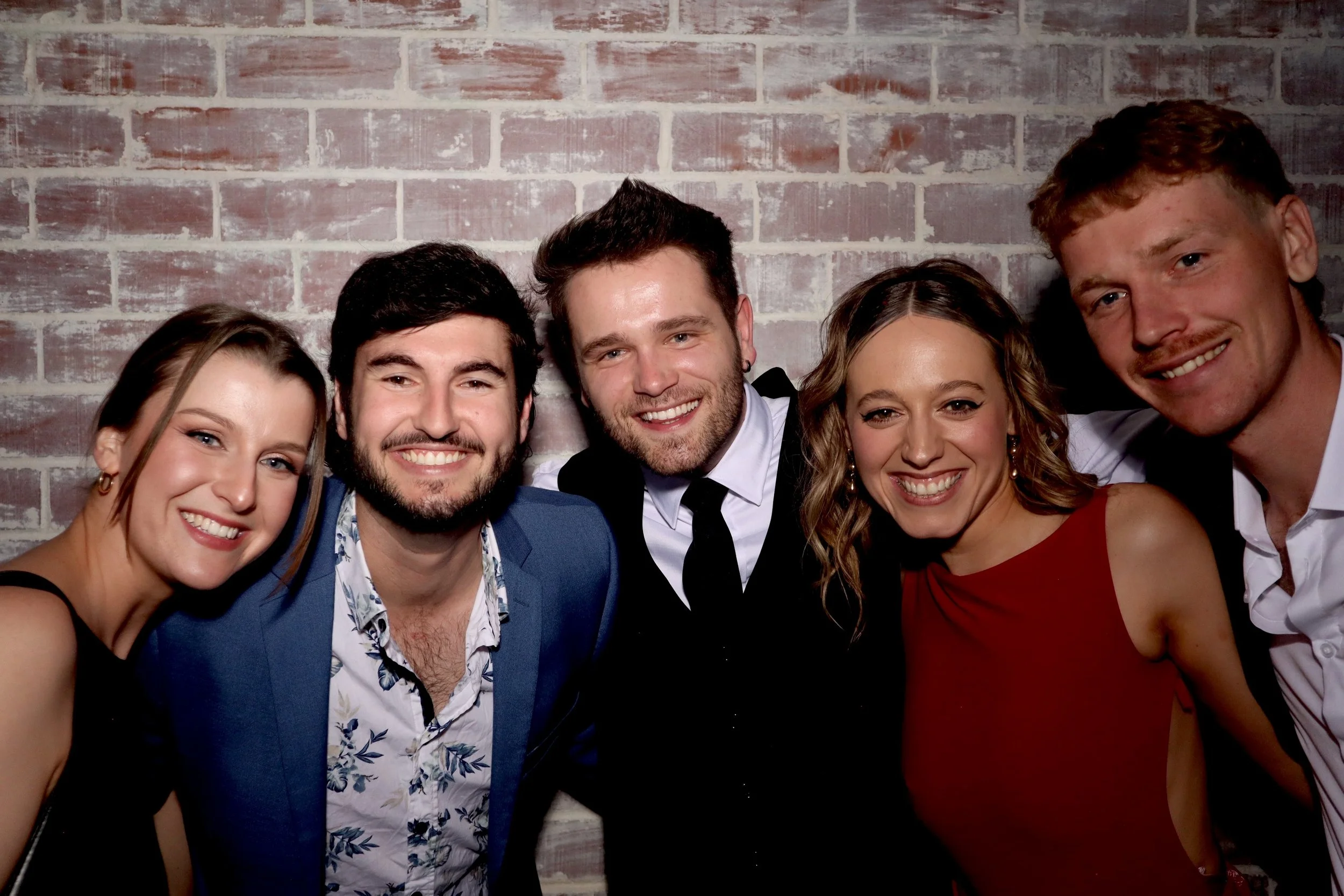 Group of five young adults smiling and posing together indoors against a brick wall. The group includes two women and three men, dressed stylishly for a social event.