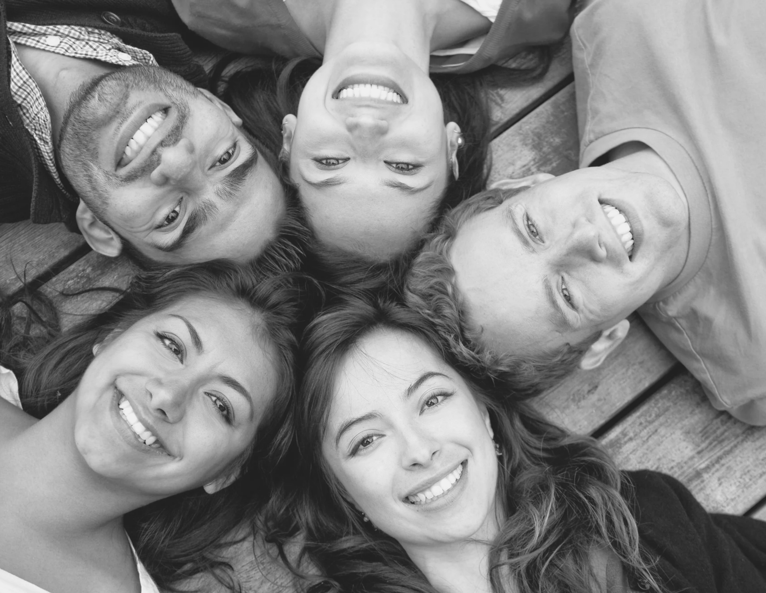 Group of people lying on the ground in a circle, smiling and looking up at the camera. Black and white photo.