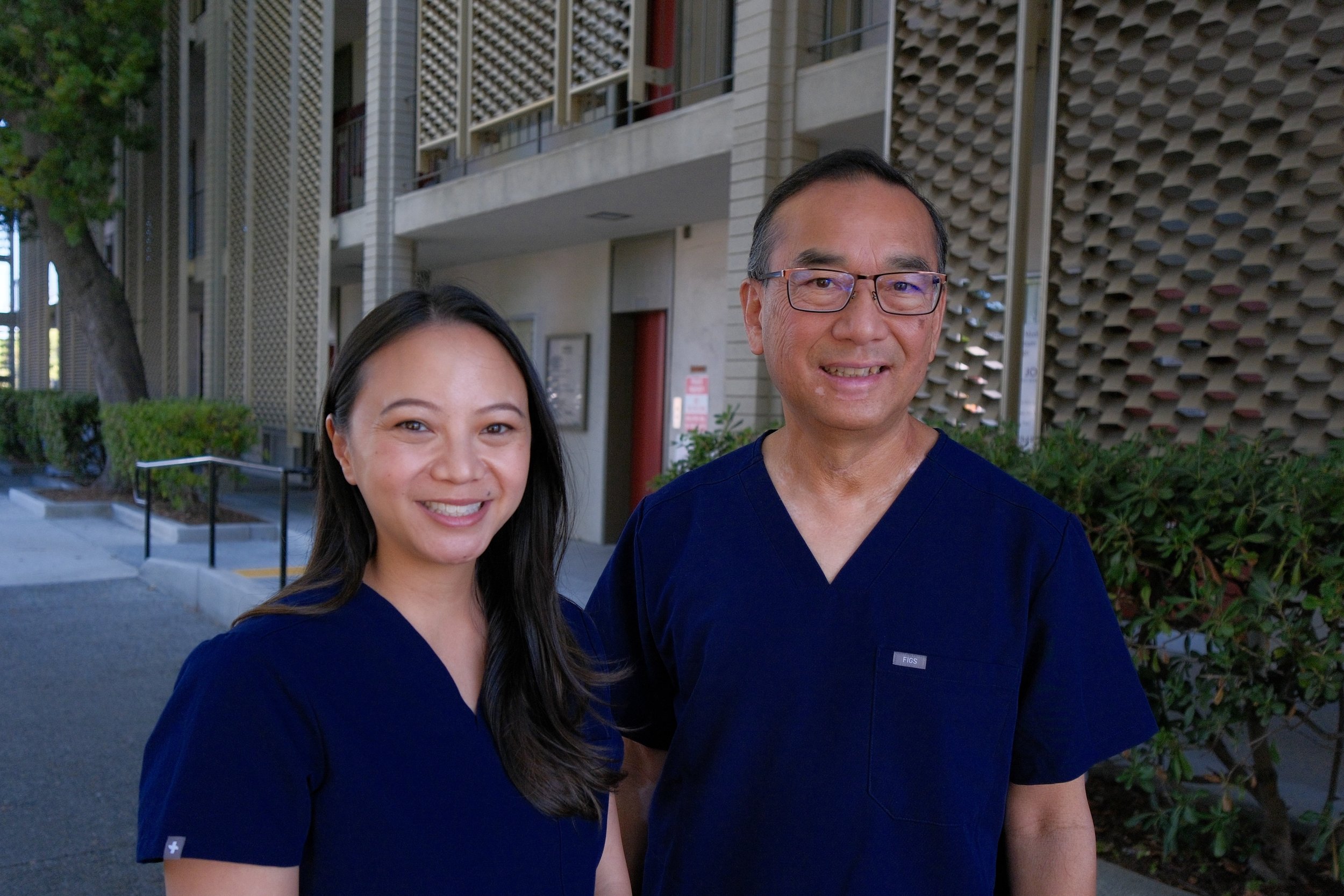 Two healthcare professionals, a woman and a man, wearing dark scrubs, standing outdoors near a building with modern architecture and greenery.