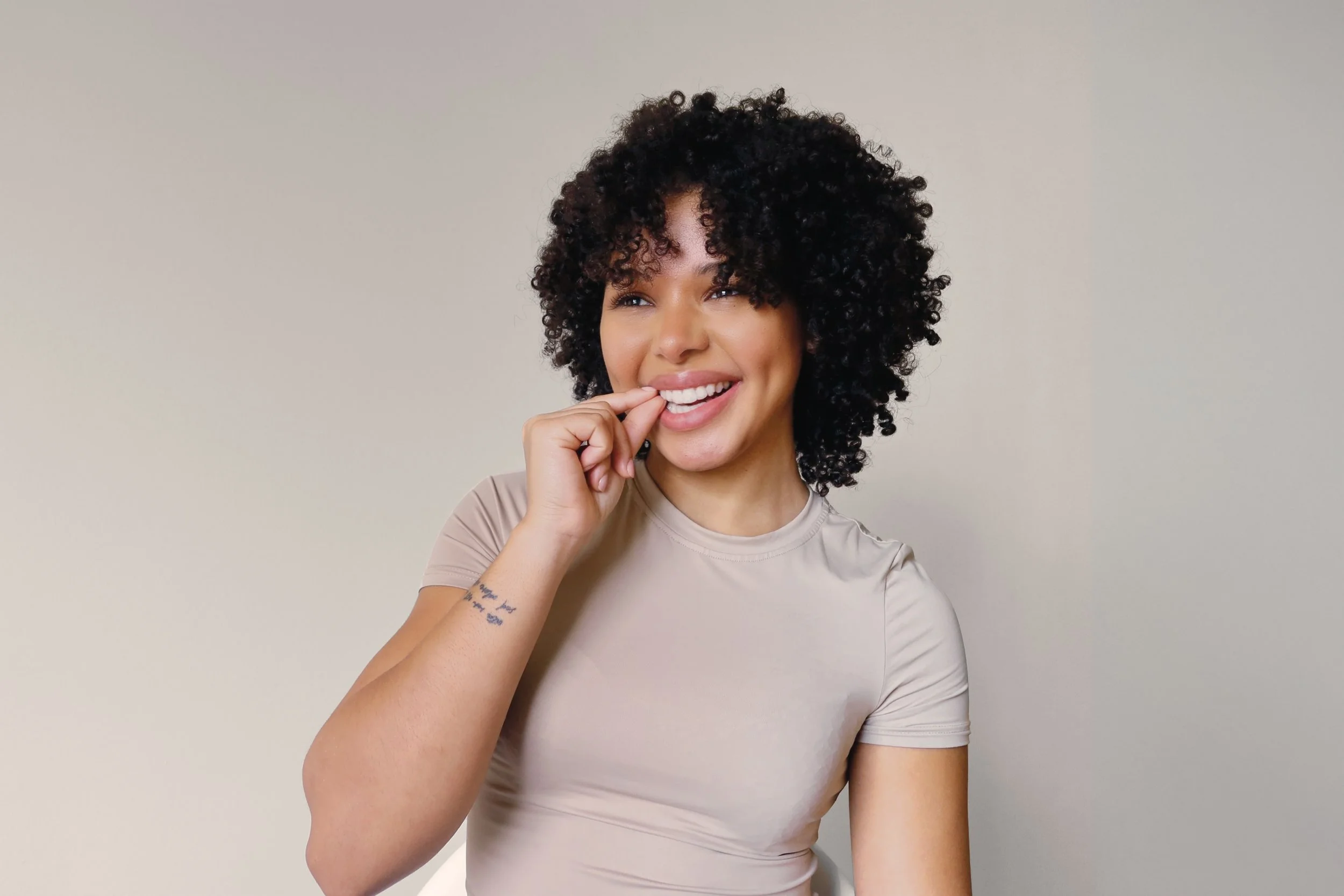 A woman with dark, curly hair smiling and biting her finger, wearing a beige t-shirt, standing against a plain background.