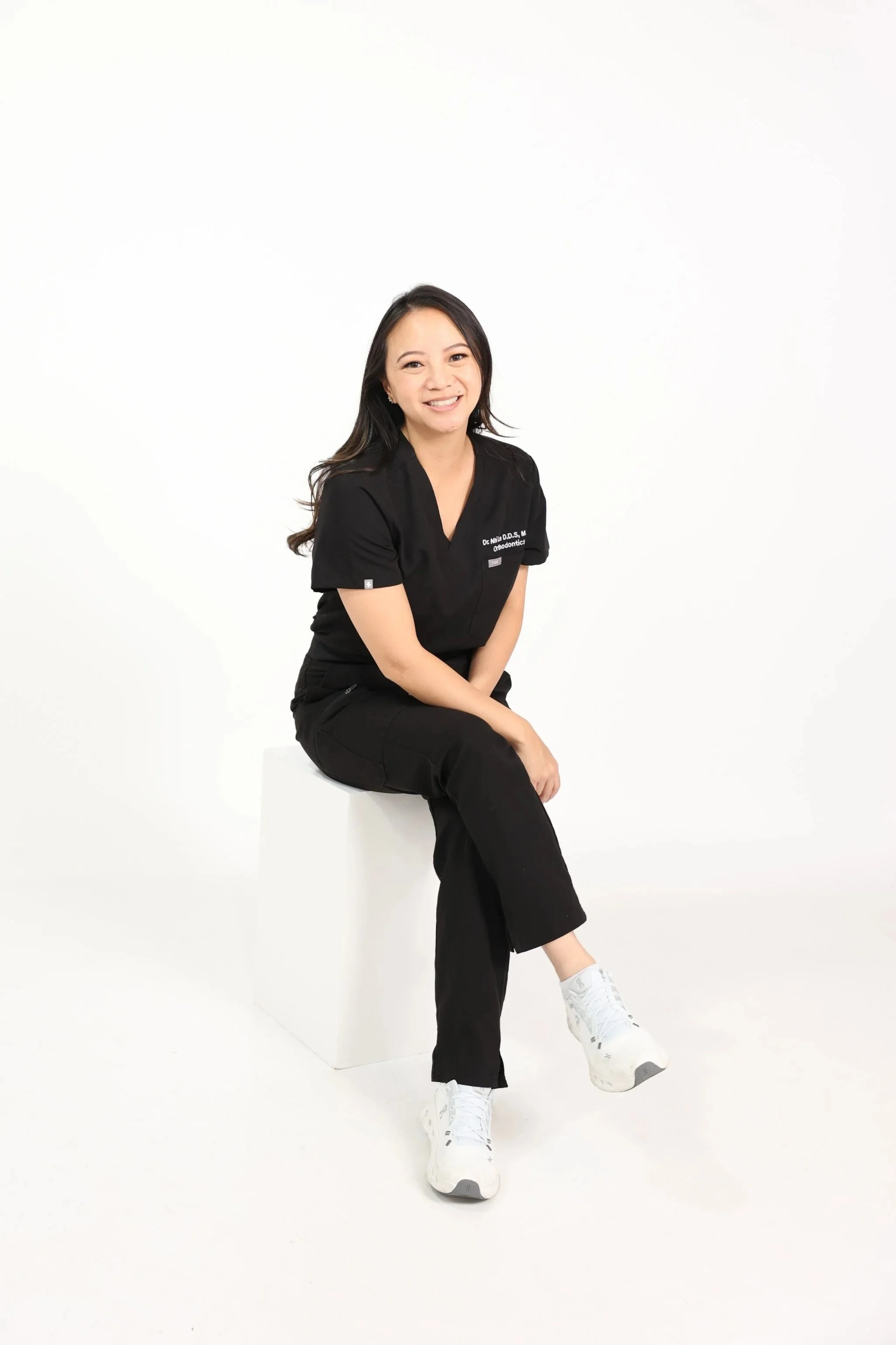 A woman sitting on a white cube against a white background. She is wearing a black professional outfit with white sneakers, smiling, with long dark hair.