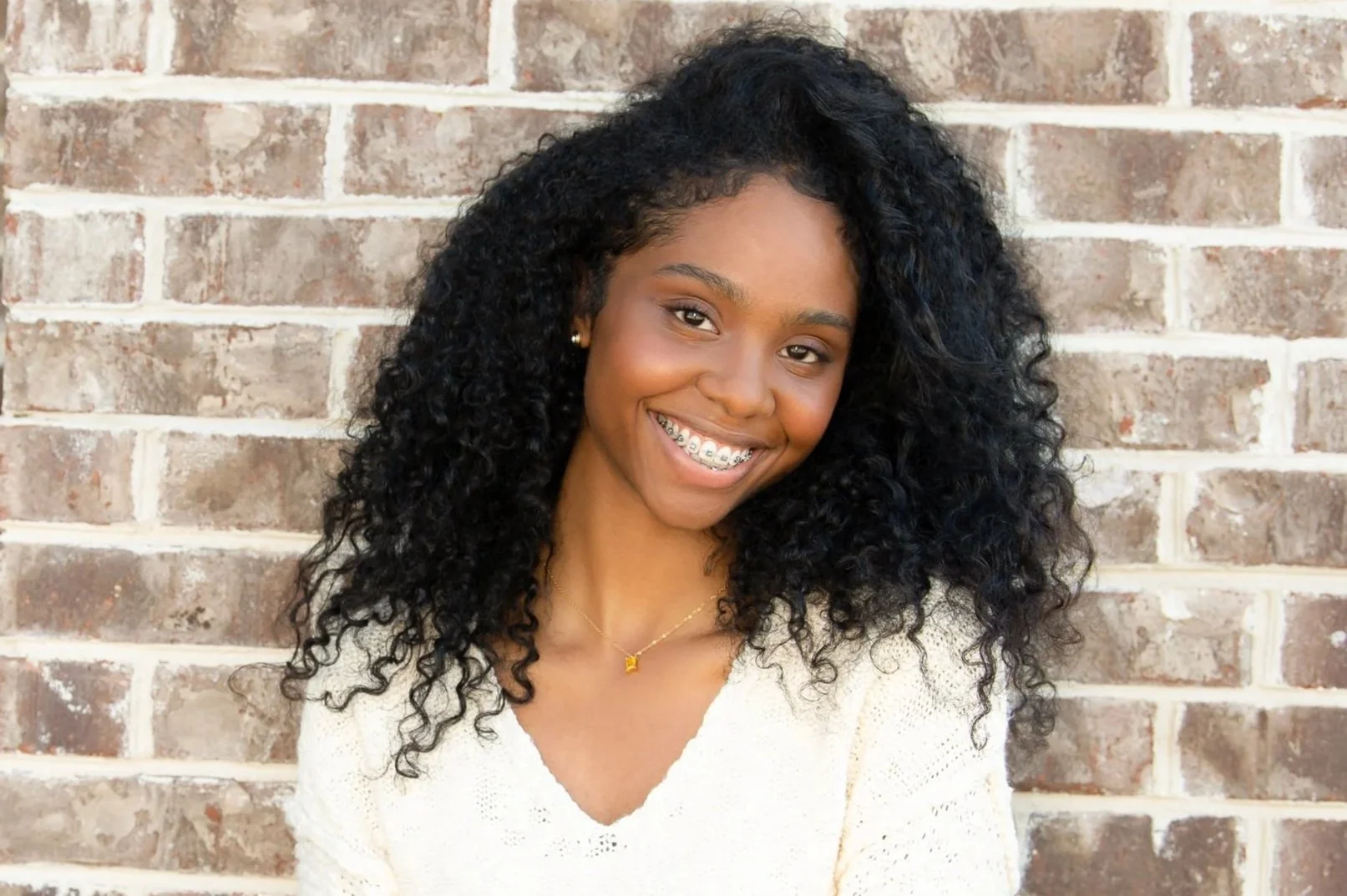 A young woman with curly black hair smiling in front of a brick wall. She is wearing braces, a white sweater, and a gold necklace with a small pendant.