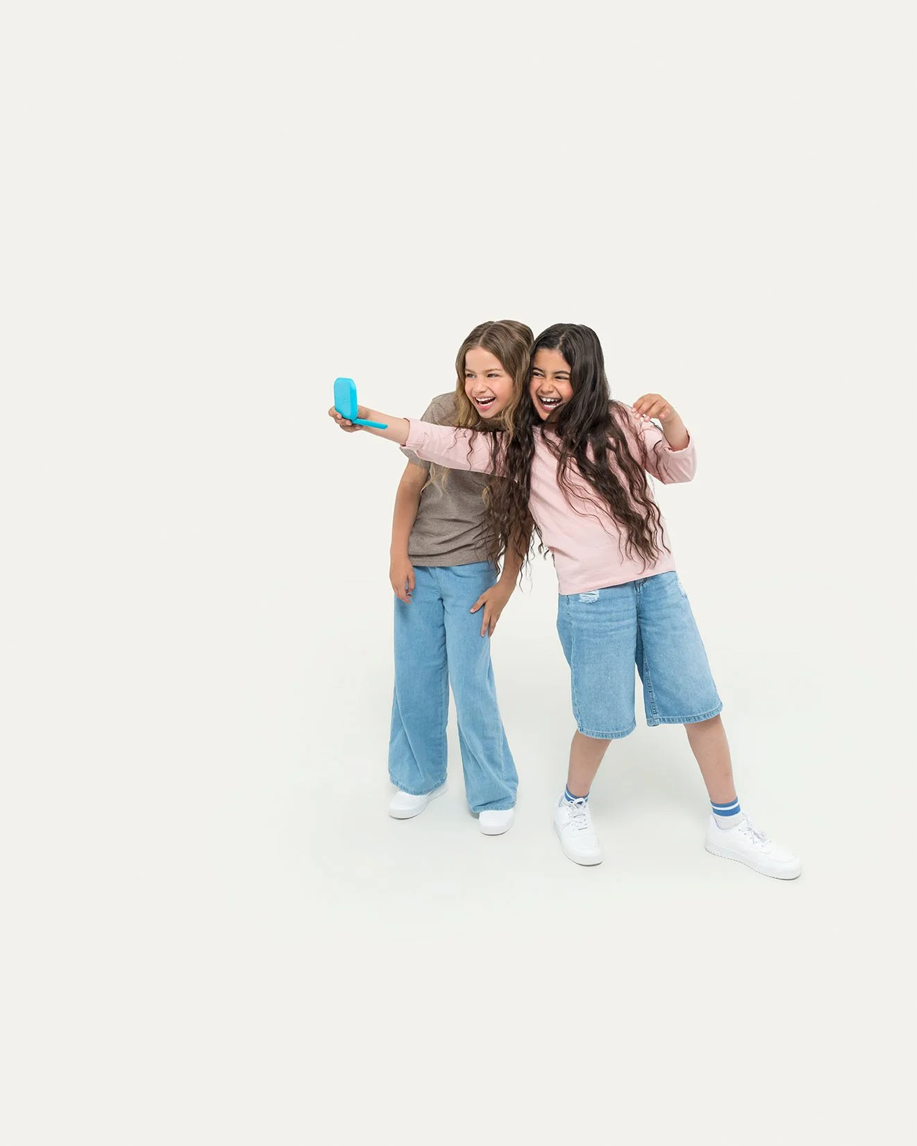 Two young girls taking a selfie together, smiling and hugging, in casual clothing against a plain white background.